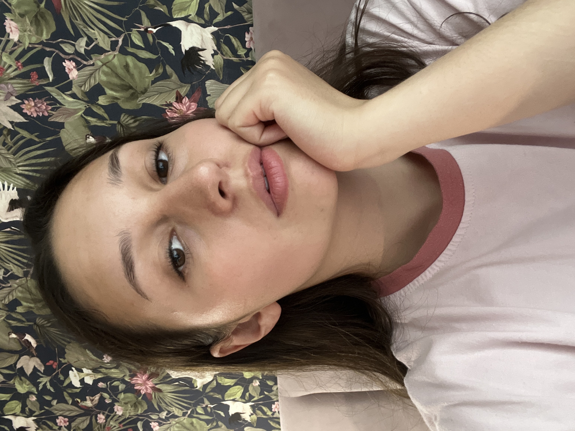 A young woman with long, dark hair is captured in a close-up, selfie-style shot. She is resting her right hand against her cheek, looking directly at the camera. Her expression is neutral, with her lips slightly parted. She is wearing a light pink t-shirt with a darker pink trim around the collar. The background features a dark, floral wallpaper with green leaves and pink flowers, and two cranes. The lighting is soft, illuminating her face. The image appears to have been taken indoors.