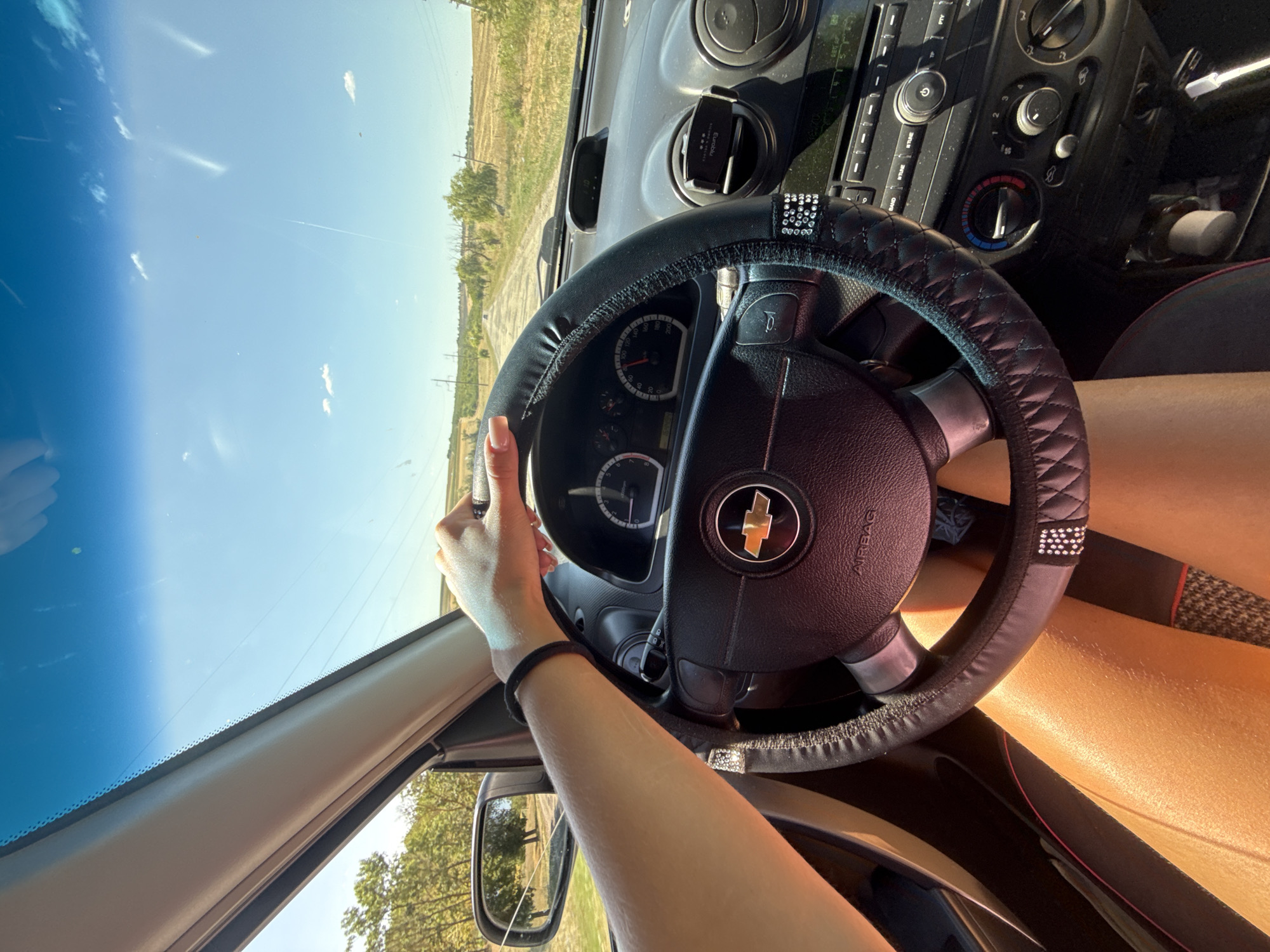 The image depicts the interior of a car as seen from the driver's perspective. A person's arm and hand, adorned with a black bracelet and rings, are visible on the steering wheel, which has a black cover embellished with rhinestones. The dashboard displays various controls, including air conditioning dials and a radio. Through the windshield, a dirt road stretches into the distance, flanked by fields and trees under a clear blue sky with scattered clouds. The scene appears to be set in a rural area outside of Basarabeasca, Moldova, judging by the landscape. The sunlight suggests it is daytime.