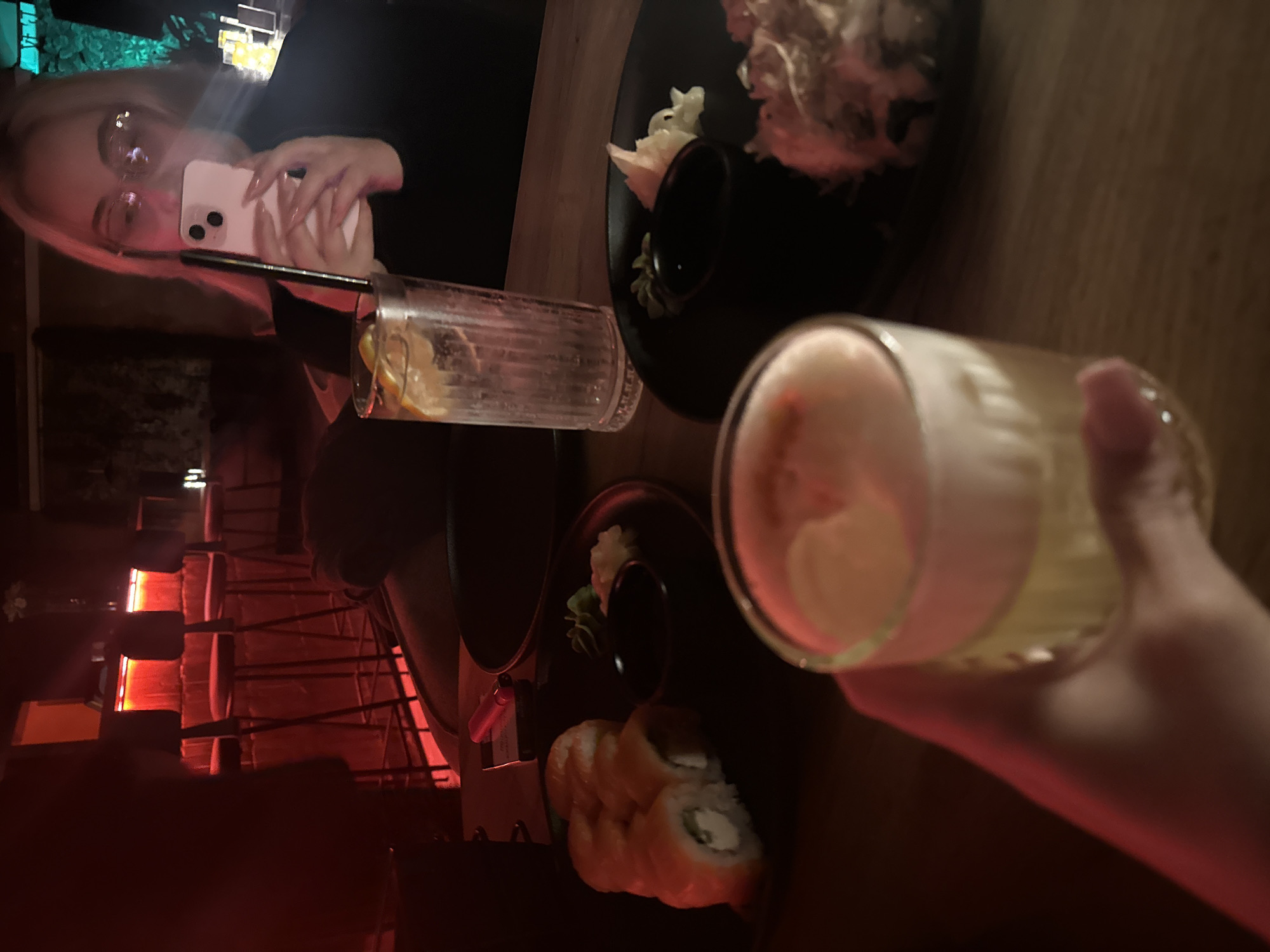 A restaurant interior in Batumi, Georgia, featuring a blonde woman taking a photo with her iPhone of a table with sushi and two cocktails. One cocktail is in her hand, while the other is on the table next to a plate of sushi. The lighting is dim, with a red hue emanating from a heater. There are several chairs visible in the background.