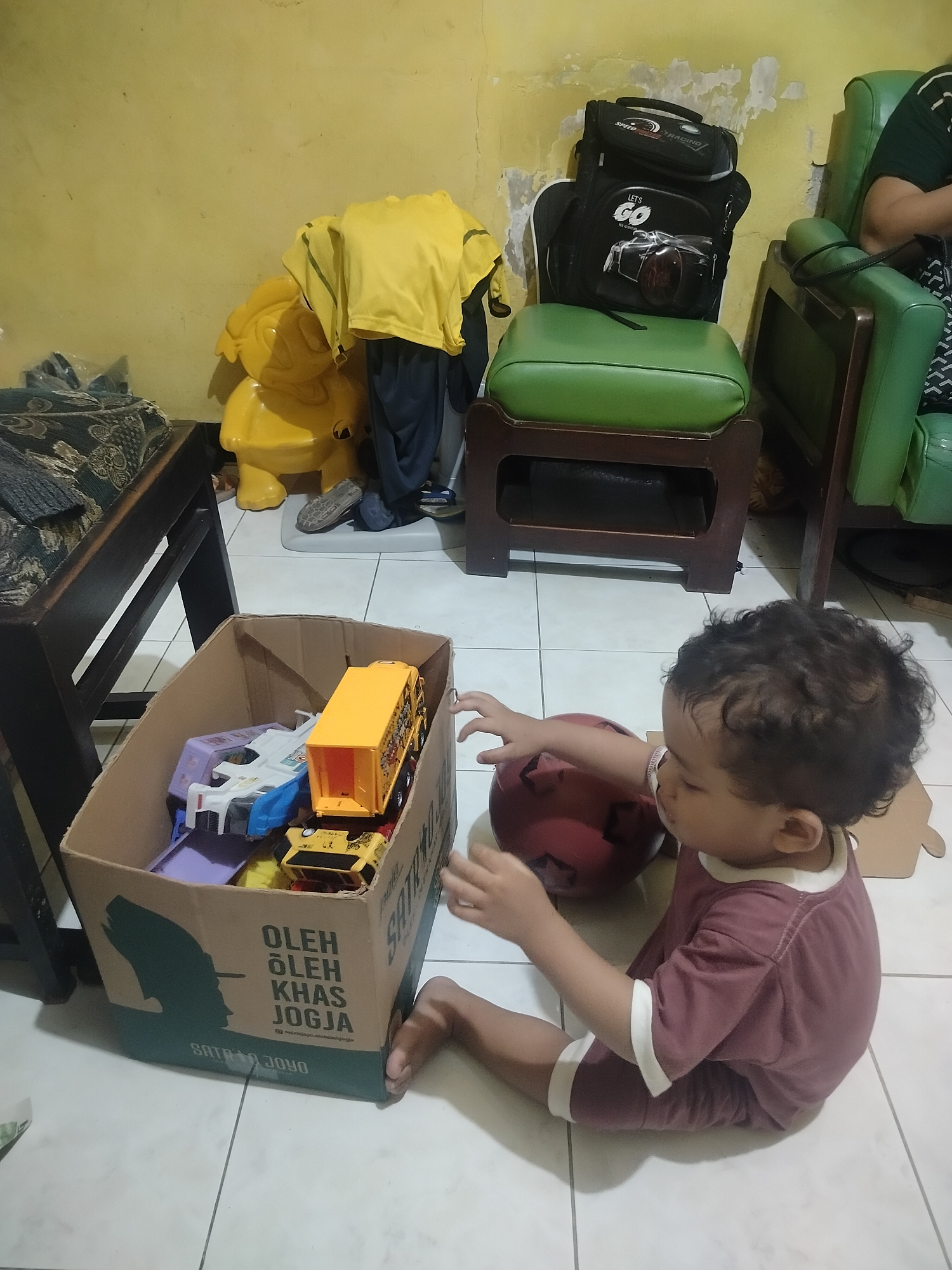 The image depicts an indoor scene within a residence in Surabaya, Indonesia. A young child, appearing to be between one and two years old, is seated on the floor. The child has dark, curly hair and is wearing a maroon t-shirt with white trim and matching shorts. The child's right hand is extended towards a red ball, and their left hand is resting on the ball as well, suggesting engagement with the toy. In the foreground, a cardboard box filled with toy vehicles, including a yellow truck, is visible. The box has text printed on its side. The floor is tiled with white squares. In the background, a yellow plastic toy, a green chair with a backpack on it, and a dark wooden table are present. A portion of another green chair is also visible. The walls are painted a light yellow, with some visible texture and discoloration. The overall impression is of a casual domestic setting where a child is playing with toys.