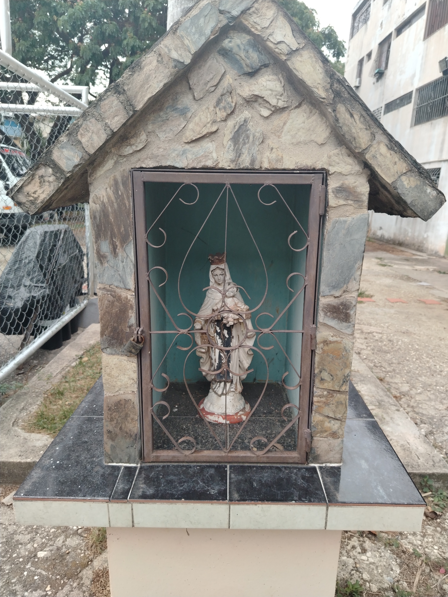 A small, weathered shrine stands outdoors in what appears to be Tinaquillo, Venezuela, based on contextual information. The shrine is constructed of stone and has a peaked roof. A metal gate with decorative curls secures the shrine. Inside is a statue of the Virgin Mary holding the baby Jesus. The statue shows signs of age and weathering. The interior walls of the shrine are painted blue. The base of the shrine is tiled with black and white tiles. The area around the shrine is paved, with some greenery visible. In the background, there is a chain link fence, a vehicle, and a multi-story building, suggesting an urban or semi-urban setting. No people are visible in the image.