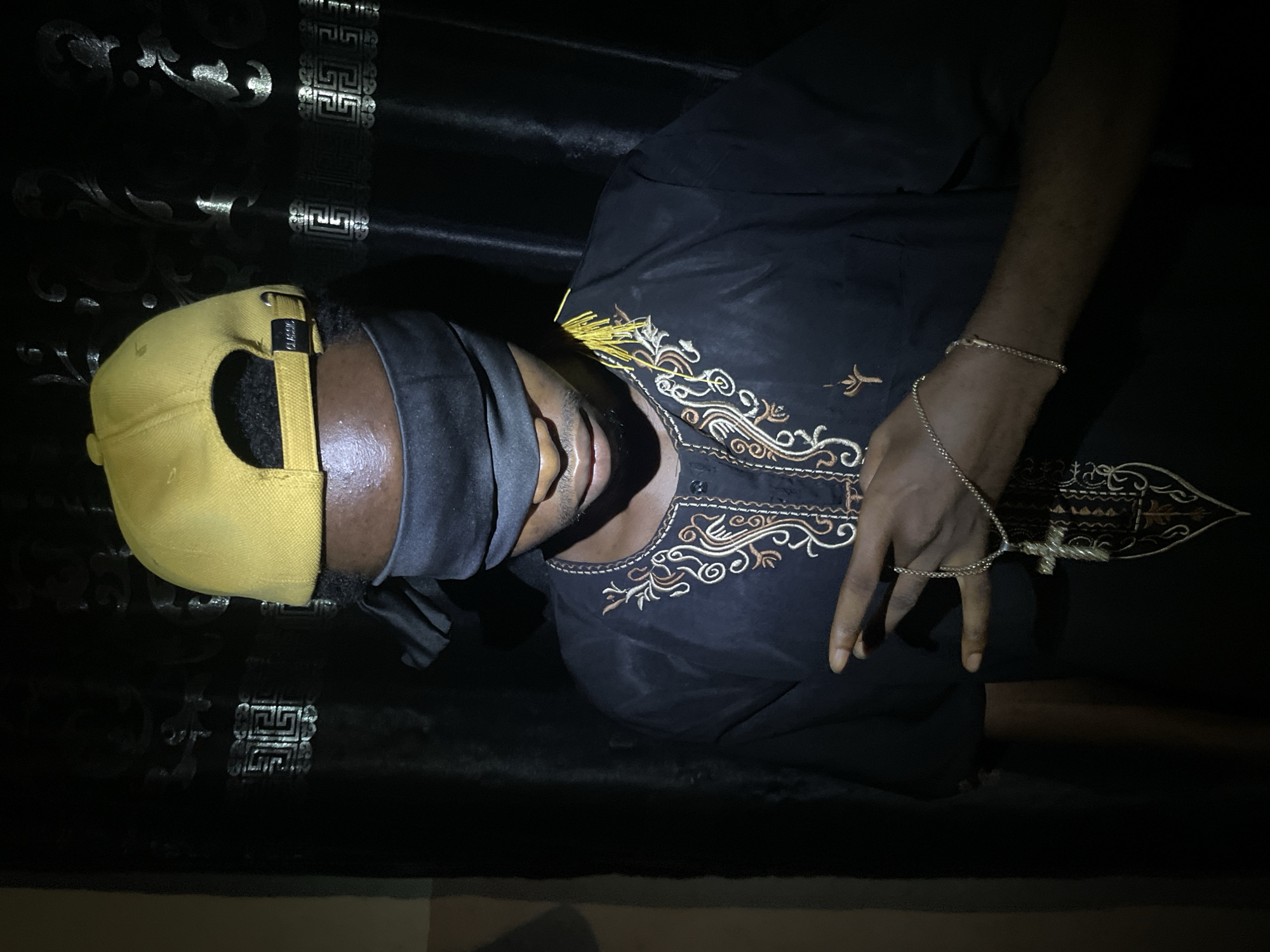 A person wearing a yellow cap and a black eye mask lies on a dark surface with decorative patterns. The person is wearing a black shirt with ornamental embroidery around the neckline and is holding a cross pendant attached to a ring on their middle finger, with a matching bracelet on their wrist. The setting is dark and difficult to discern with details about the location in Lokoja, Nigeria being indeterminate from the image.