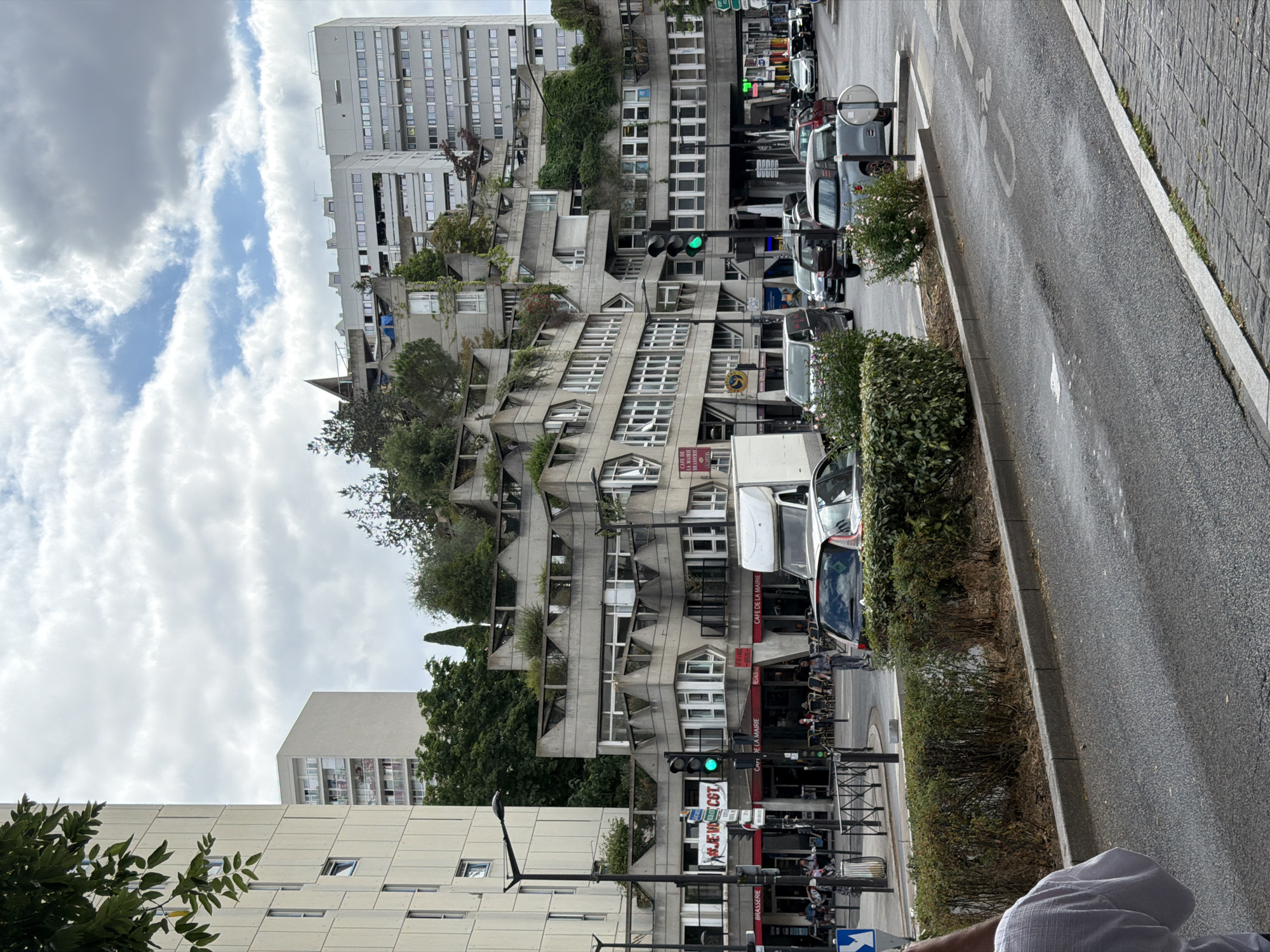 The image captures a street scene in Ivry-sur-Seine, France, focusing on a distinctive Brutalist-style building complex. The building features a tiered, concrete facade with geometric shapes, extensive balconies, and integrated greenery, giving it a terraced, almost organic appearance. In the foreground, a two-lane asphalt road runs horizontally across the frame, marked with a white bicycle lane symbol. Cars are visible on the road, including a dark-colored hatchback in the right lane and other vehicles further down the street. A white delivery truck is parked on the right side of the road. On the left side of the street, a sidewalk with a metal railing separates pedestrians from the road. Several pedestrians are seated at outdoor tables belonging to a brasserie or cafe with red awnings. A traffic light displaying a green signal is visible. Behind the Brutalist building, a tall, modern, white apartment building with numerous windows stands prominently against a cloudy sky. Trees and vegetation are interspersed throughout the scene, adding a natural element to the urban environment.