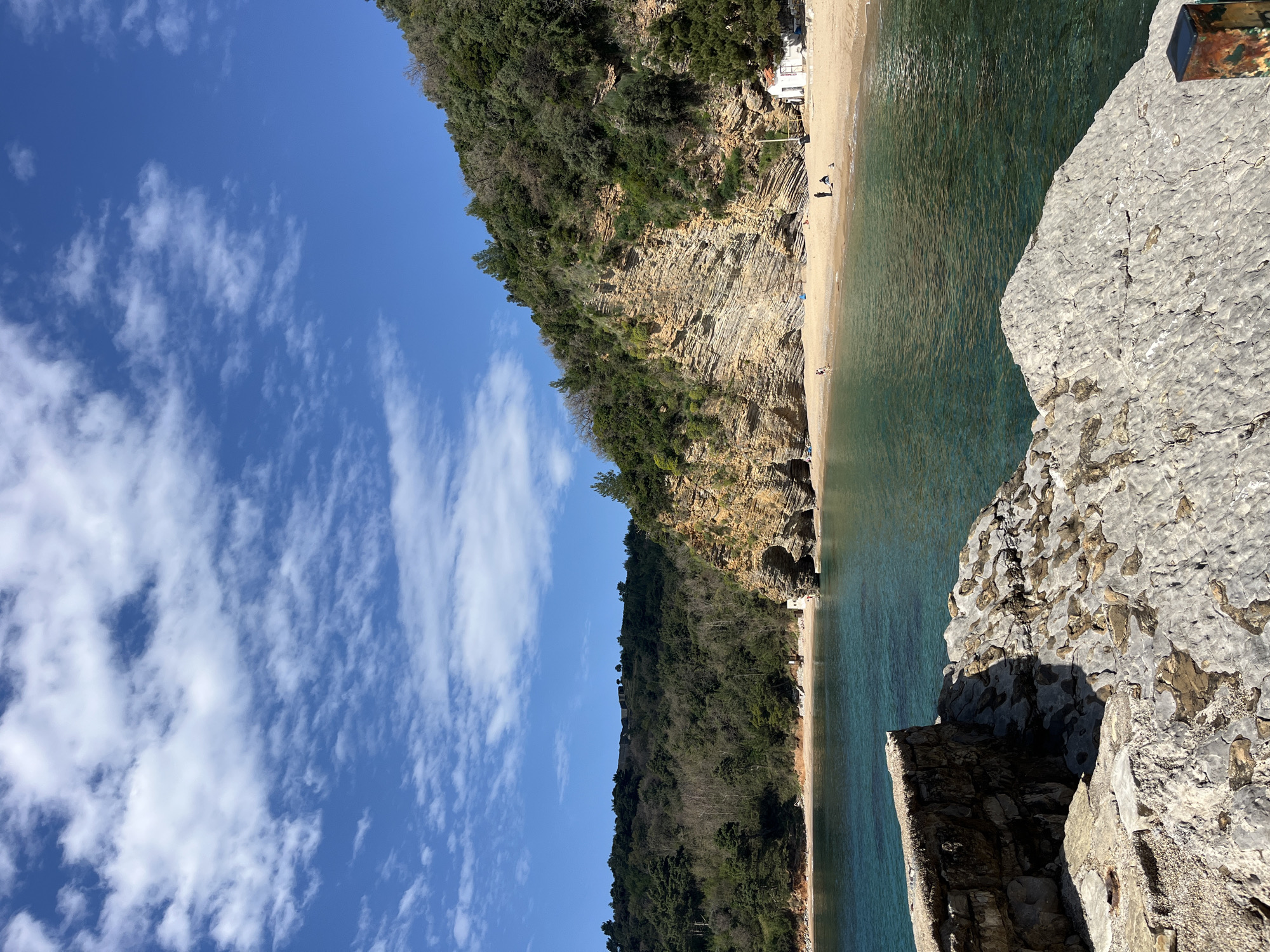 Here is a description of the image:
The image presents a landscape view, oriented sideways, showcasing a beach setting adjacent to a rocky terrain with lush vegetation under a partly cloudy, blue sky.
**Location:** The scene appears to be located in or near Budva, Montenegro.
**Scene Details:**
* **Beach:** A narrow sandy beach is situated along the coastline. Several people are seen on the beach.
* **Water:** The water transitions from turquoise in shallower areas to a deeper green further out.
* **Cliff/Vegetation:** A cliff or a rocky elevation is visible, covered with green vegetation, likely trees and shrubs. The cliff has visible geological strata.
* **Sky:** The sky is blue with scattered, puffy white clouds.
* **Foreground:** A rocky structure is visible in the foreground, indicating the vantage point of the photograph. The rocky formation appears weathered, with patches of discoloration.
* **Structure:** A white structure is at the base of the cliff near the water.
The environment is typical of the Adriatic coast, with clear waters, rocky terrain, and Mediterranean vegetation.