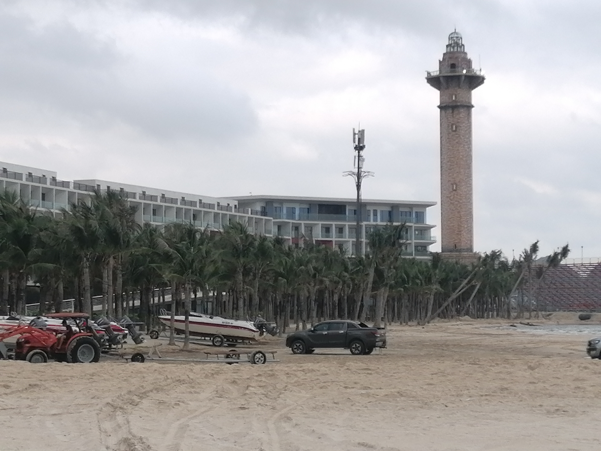 This wide shot captures a coastal scene in Bai Chay Ward, Vietnam, under a uniformly overcast sky. The foreground is dominated by a light brown sandy beach, showing various tire tracks and disturbed patterns.
In the midground, several objects and activities are visible. On the left, a red tractor is positioned on the sand, adjacent to multiple white speedboats resting on trailers. Further to the right, a dark-colored pickup truck is parked on the sand, near more boats on trailers. Behind these vehicles and boats, a dense row of palm trees stretches horizontally across the image. Many of these trees, especially towards the right, appear to be leaning significantly, suggesting exposure to wind or a natural growth pattern.
The background features several structures. A long, modern, multi-story building, likely a hotel or resort, with numerous balconies and light-colored facades, occupies the left and central parts of the upper frame. To the right of this building, a tall, slender, reddish-brown lighthouse-style tower with an ornate top section stands prominently. Between the hotel and the lighthouse, a tall cellular or communication tower with antennas is visible. On the far right, beyond the palm trees, a section of a large, tiered seating structure, possibly bleachers or an amphitheater, can be seen.
The time of day is daylight, but the heavy overcast clouds create a soft, diffused light, indicating a cloudy or potentially rainy day. No specific activity involving people is clearly depicted, though the arrangement of boats, trailers, and the tractor suggests boat launching, retrieval, or storage operations. No discernible text is visible on any objects or structures. The overall scene conveys a functional coastal area with resort amenities.