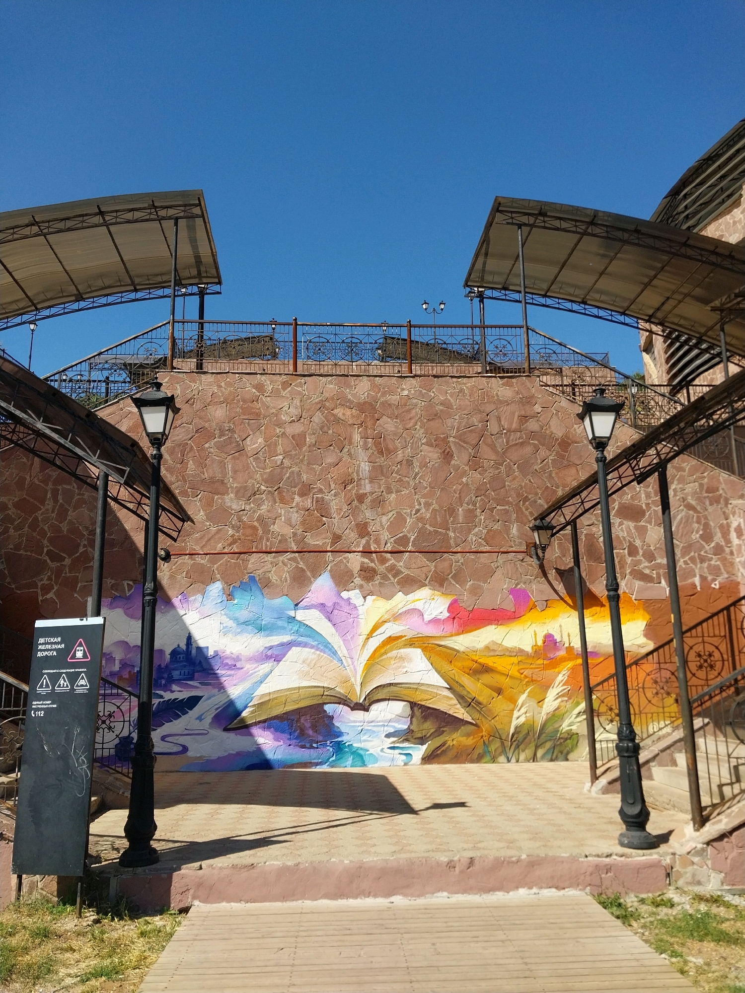 This is a photograph of a mural depicting an open book with pages flowing into a cityscape and natural elements, located on the exterior wall of a building. The mural is vibrantly colored with blues, purples, yellows, and oranges. Above the mural, the stone wall continues upwards, topped with a metal railing. On either side of the central wall are staircases with metal railings, leading up to a higher level. The staircases are covered by awnings supported by metal poles. Two black lampposts are visible in the foreground. To the left of the mural, a black sign with white text and symbols is mounted. The ground in front of the mural is paved with interlocking square tiles, and a wooden walkway is in the foreground. The sky is clear and blue. The scene is bathed in sunlight, casting shadows. This mural is located in the city of Orenburg, Russia, near a children's railway.