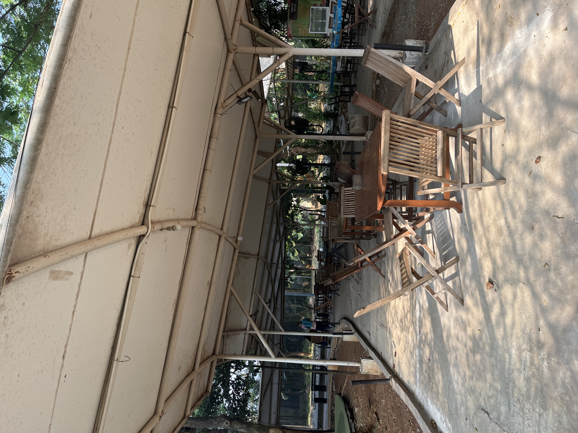This image depicts an outdoor seating area under a large, light-colored canopy, likely at a cafe or restaurant in East Jakarta, Indonesia. The seating area is comprised of several wooden tables and chairs arranged on a concrete patio. In the foreground, a wooden table with slatted chairs is prominently featured, with shadows from the canopy and surrounding trees dappling the ground.
Further into the scene, more tables and chairs are visible, some with patrons. A person in a patterned shirt and dark pants is walking away from the camera, further into the covered area. To the left, there is a glimpse of a grassy area and some dirt landscaping. The background reveals dense foliage and trees, suggesting a location with ample greenery. The overall impression is of a relaxed, open-air dining or resting spot.
