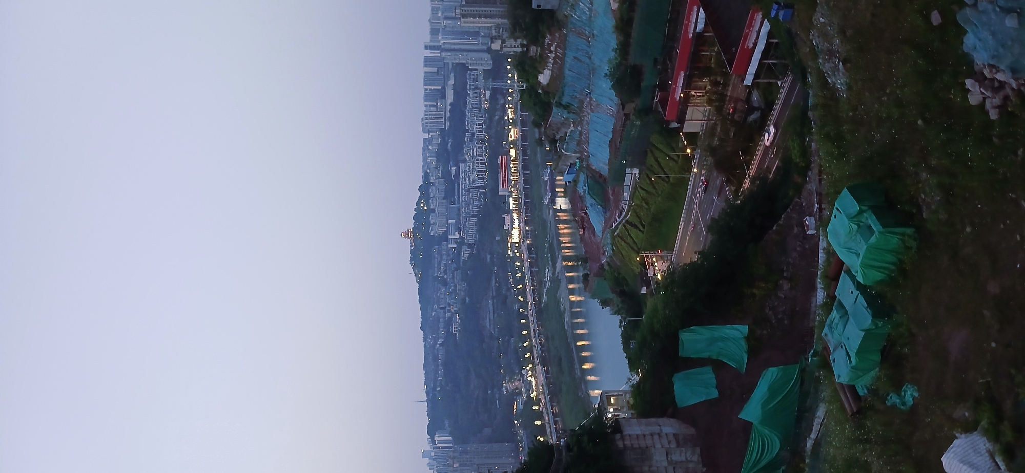 This is an outdoor, elevated shot of a cityscape, taken around dusk or dawn. The sky is a pale blueish-gray. A dense collection of buildings, illuminated with artificial lights, extends along a hillside. A river or wide waterway reflects the lights, creating a parallel line of illumination. A road, also illuminated by streetlights, runs alongside the water. On the right side of the image, there are signs of construction or other activity. There are green tarp covers over objects on a grassy area in the foreground. A building with a red roof is visible. The shot is consistent with an image taken in or around the Yuzhong District of Chongqing, China, based on the type of architecture and density of buildings. There are no visible people in the image. There is moderate traffic visible on the road.