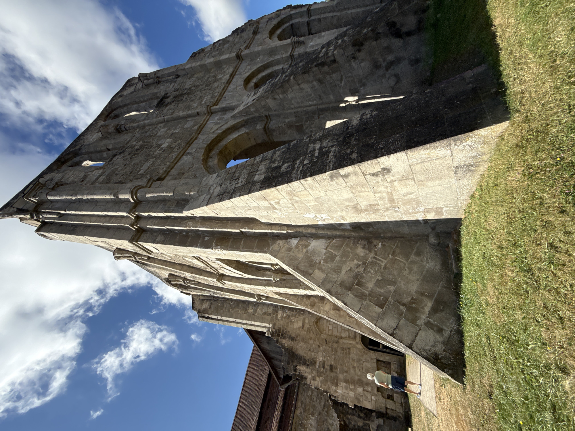 An upward, wide-angle shot captures the imposing, weathered stone ruins of the Walkenried Abbey, a historic Cistercian monastery located in the municipality of Walkenried, Germany. The sky above is a vivid blue, dotted with white, wispy clouds. The ruins, constructed from large, rough-hewn stone blocks, feature towering walls with arched openings, showcasing the architectural remnants of a grand structure. In the foreground, lush green grass covers the ground, transitioning to drier, yellowish vegetation near the base of the ruins. A single person, seen from the back, stands on a paved path to the left of the ruins, appearing to observe the ancient edifice. The person is wearing a light-colored t-shirt and dark shorts. The sun casts strong shadows, highlighting the textures and depth of the stone masonry.