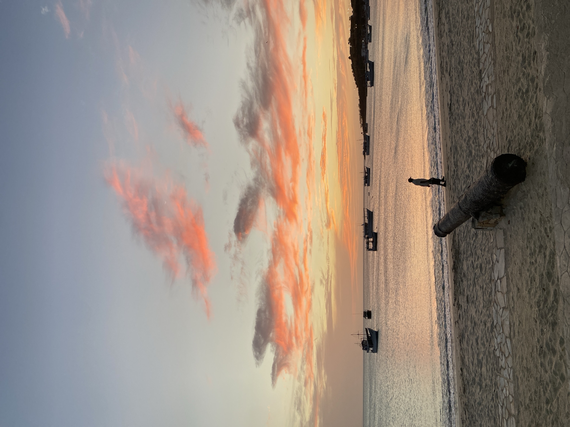 The image shows a sunset over the ocean in Juan Griego, Venezuela. The sky is a mix of blue, orange, and pink, with clouds streaked with orange and gray. Several boats are anchored in the calm water. A person stands on the beach near the water's edge. A cannon rests on a stone wall that borders the beach. The beach itself appears to have a narrow strip of sand before the low wall. The location is likely near the Fortin de La Galera, based on the cannon and seaside setting.