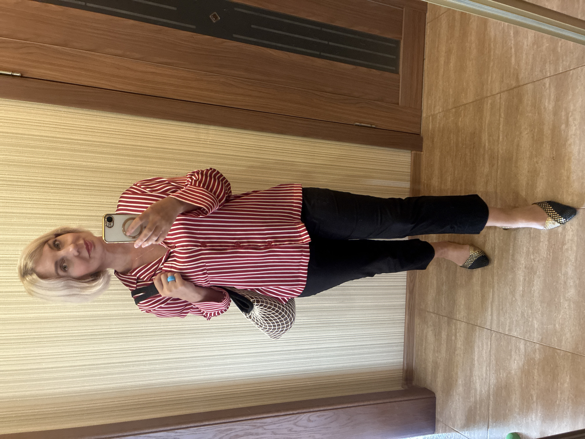 A woman is taking a mirror selfie in what appears to be a room with striped wallpaper and tiled flooring. She has short, blonde hair and is wearing a red and white striped button-down shirt and black pants. She is also wearing black and gold patterned flat shoes. She is holding a smartphone in her left hand and has a turquoise ring on her right ring finger. A mesh handbag is tucked under her left arm. The background includes a wooden door frame and a reflective surface that shows a portion of the room and the woman. The lighting suggests it is daytime. The location is identified as Sochi, Russia. There is no visible text in the image.