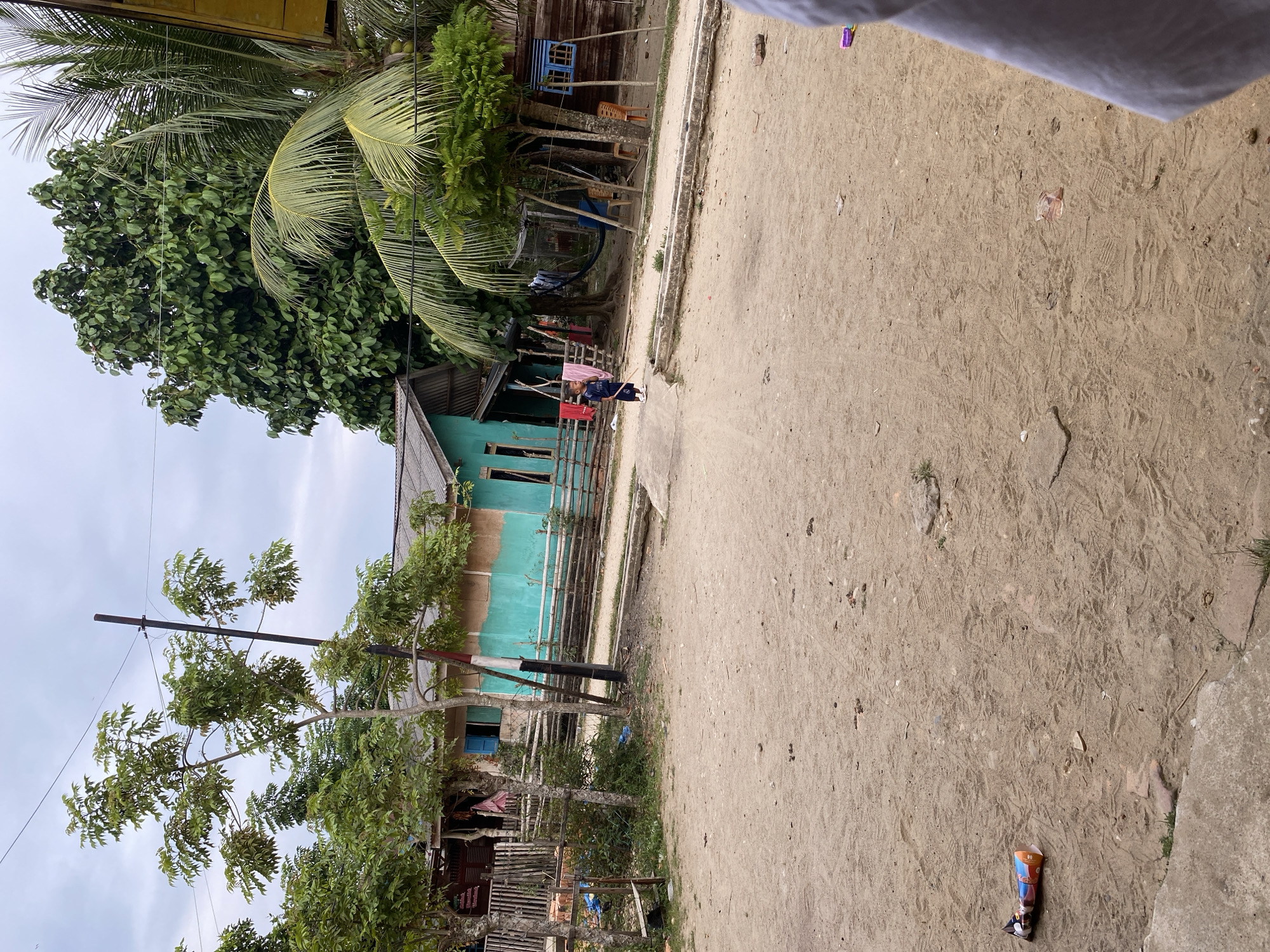 This image captures a street scene in Tanjongkapai, Indonesia, on an overcast daytime. The perspective is slightly elevated, looking across a wide, sandy dirt path that occupies most of the right side of the frame. The path is marked with numerous footprints and scattered small debris.
On the left side, the scene is dominated by lush tropical vegetation, including large palm trees with visible coconuts and other dense, leafy trees. Several houses are visible amongst the greenery. A prominent, two-story house features a distinctive turquoise facade with blue window frames. Further back, another wooden house is partially obscured. Simple wooden and bamboo fences delineate the property lines. Clothes are seen hanging to dry near one of the houses, suggesting daily life. A black utility pole with visible wires stands tall, indicating electrical or communication infrastructure.
In the mid-ground, a young person, likely a child, dressed in dark clothing, is walking along the dirt path. In the immediate foreground, on the path, a colorful, crinkled snack wrapper is discarded. The sky overhead is a uniform grey, indicating cloudy weather. The overall setting is that of a rural or semi-rural residential area, characterized by unpaved roads, abundant vegetation, and simple dwellings. No specific legible text is visible, only unidentifiable graphics on the snack wrapper.