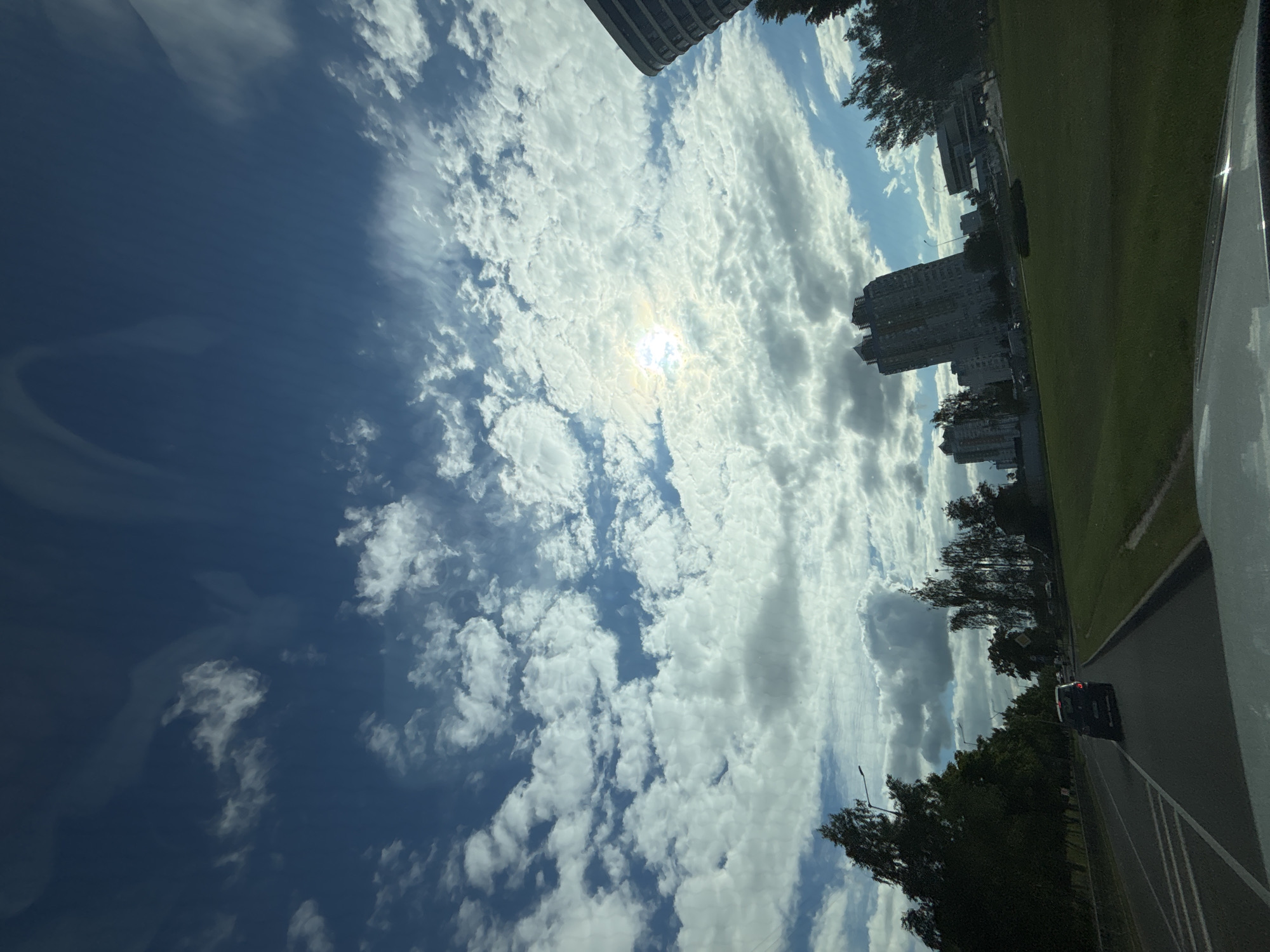 The image depicts a scene from a moving vehicle, likely on a road within the city of Minsk, Belarus. The sky is a prominent feature, filled with a mix of white cumulus clouds and thinner, wispy cirrus clouds, with the sun peeking through, creating a bright glare. Below the sky, there are several multi-story apartment buildings visible, characteristic of urban landscapes. Green grass lawns and trees line the side of the road. A dark-colored car is driving ahead of the viewer's vehicle on a multi-lane road. The perspective suggests a forward-facing view from the driver's or passenger's seat. There are no people clearly visible in the frame.