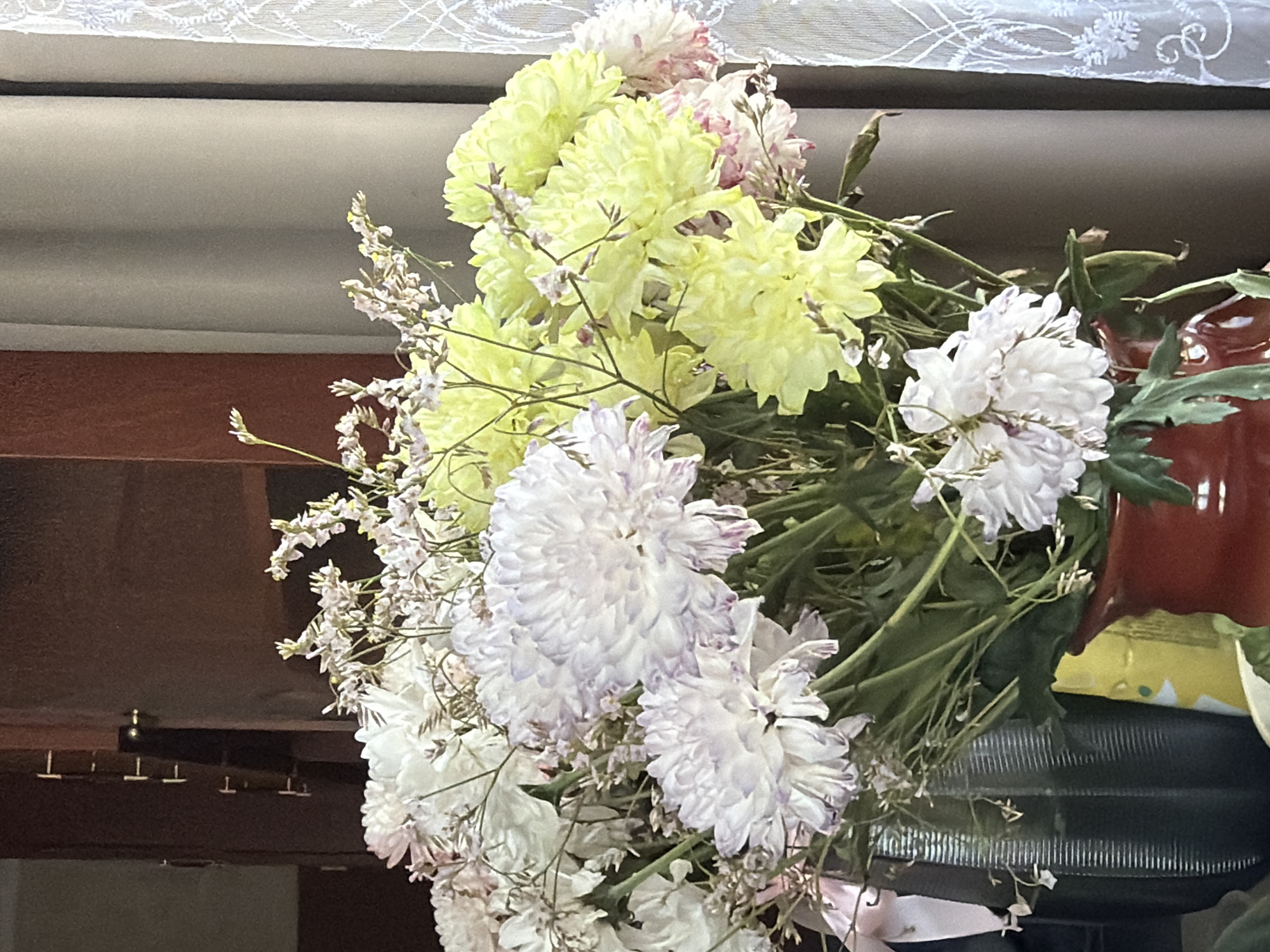 The image depicts a bouquet of flowers in an indoor setting. The bouquet contains chrysanthemums in colors of yellow and white with hints of light purple, along with smaller white filler flowers. Green stems and leaves are visible. The bouquet is positioned in front of a dark wood cabinet or surface. A white lace curtain is partially visible in the upper portion of the image. The location, Чумна, Russia, is not discernible from the visual data provided.