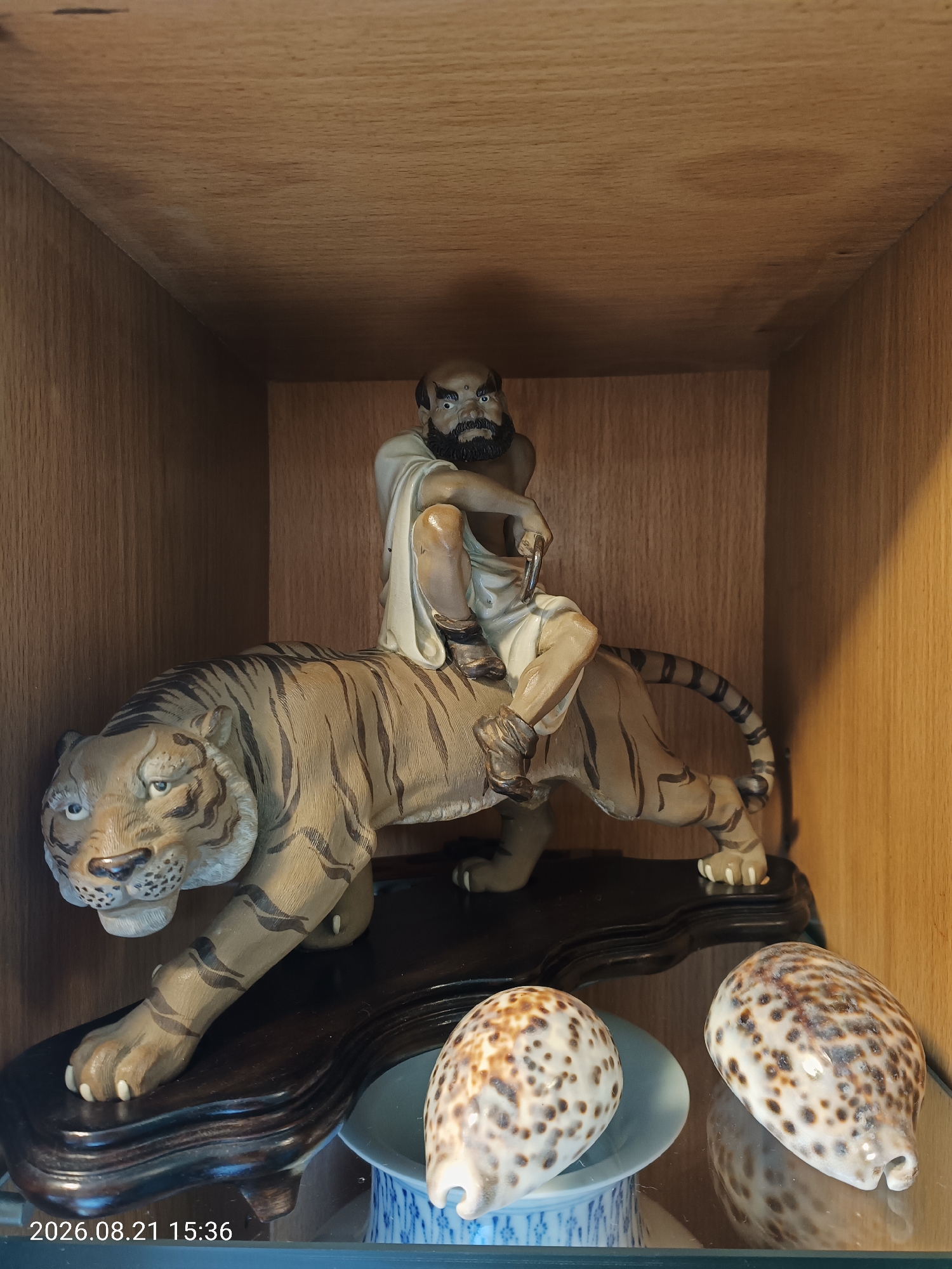 The image shows a figurine of a man riding a tiger. The figurine is placed on a wooden shelf with two seashells in the foreground. The man in the figurine has a beard and is wearing a white garment. The tiger is in a stalking pose with its mouth open. The background is a wooden shelf with vertical wooden planks. There is a timestamp in the bottom left corner of the image which reads "2026.08.21 15:36". The image does not provide any information to determine its location in Hong Kong, China.