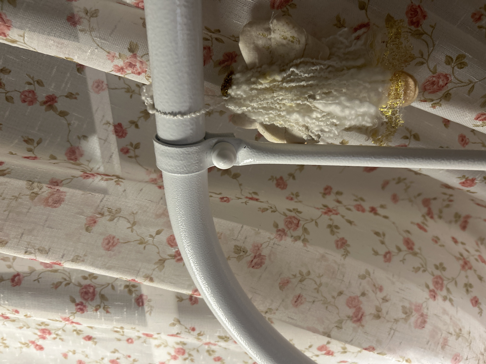The image shows a close-up of a white metal bed frame with a small angel ornament hanging from it. The ornament is made of white fabric with a fluffy white hair-like texture, and it has gold accents. Behind the bed frame are sheer curtains with a pattern of pink roses and green vines. The scene appears to be indoors, likely a bedroom. The location is identified as Троица, Russia. The image is focused on the bed frame and the ornament, with the floral curtains serving as a soft, decorative backdrop. There are no people or discernible actions taking place in the image.