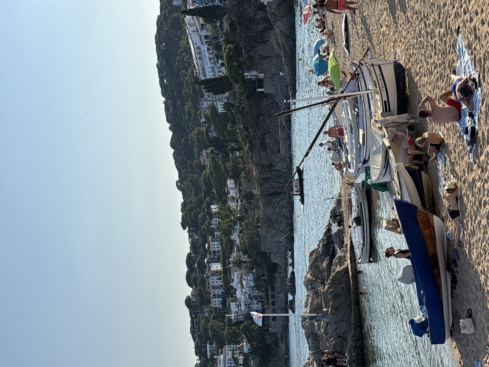 The image depicts a sunny day at Calella De Palafrugell, Spain. The scene is a beach with calm blue water and a sandy shore. Several boats are pulled up onto the beach, with some people relaxing on towels and sunbathing. In the water, a few people are swimming or wading. Further out, a larger boat with several passengers is visible. The background shows a lush green hillside dotted with white buildings, characteristic of the coastal architecture in the region. A flag is flying from a pole on a rocky outcrop in the foreground. The sky is clear and blue.