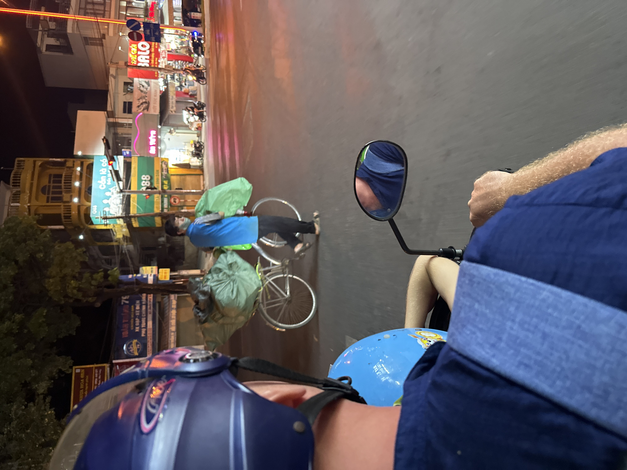A person wearing a blue helmet is riding a motorcycle at night in Bac Nha Trang Ward, Vietnam, alongside a cyclist carrying a bag. The street is illuminated by storefront signs and lights. The motorcycle's side mirror shows a reflection of the rider. The cyclist is wearing a blue shirt and dark pants. Street signs and shop fronts are visible in the background.