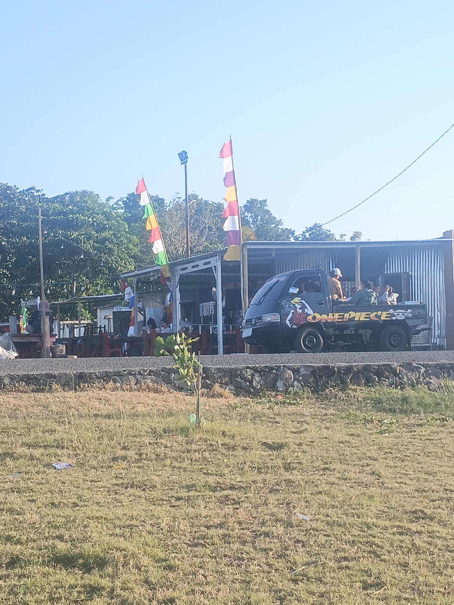 The image depicts a street scene in Lewoleba, Indonesia, under a clear blue sky. In the foreground, dry, patchy grass covers the ground, with a few pieces of litter visible. A low stone wall separates the grassy area from a road.
Dominating the middle ground is a row of low-lying structures, likely food stalls or small shops, with corrugated metal roofs supported by white metal frames. Colorful flags, predominantly red, white, and yellow, are strung between poles, adding a festive touch. Several people are seated at tables within or near these structures, appearing to be patrons or staff.
A dark-colored pickup truck, decorated with a "ONEPIECE" logo and character art, is parked in front of one of the structures. The truck appears to be carrying several passengers in its bed.
In the background, lush green trees and foliage form a dense natural barrier, hinting at the proximity of nature to the urban environment. The overall impression is a lively, casual outdoor gathering space in a tropical setting.