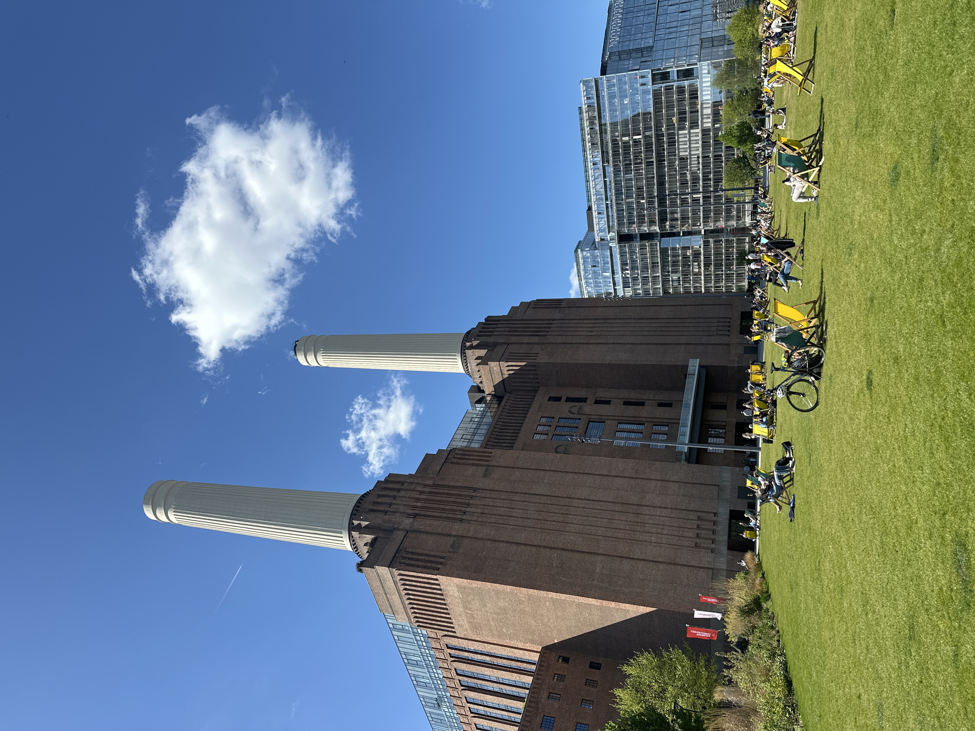 The image shows the Battersea Power Station in London, United Kingdom, on a sunny day with a clear blue sky and a few clouds. The power station's four chimneys are prominent. A modern glass building stands next to the power station. A grassy area extends towards the right side of the image, with people relaxing in chairs. A bicycle is visible among the chairs. The scene is open and public, suggesting a recreational space in an urban environment. Flags with red and white lettering are planted in front of some trees.