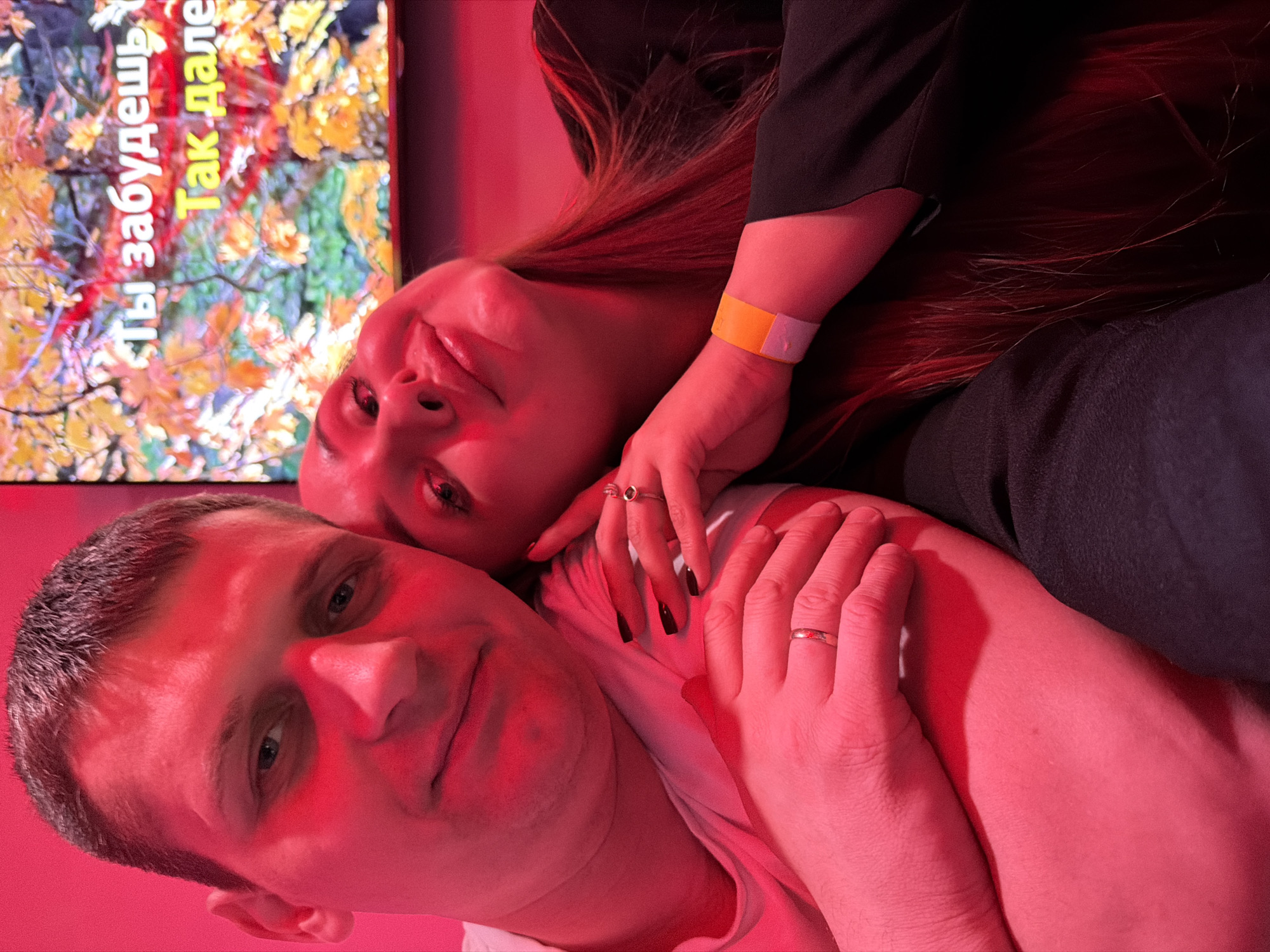 The image depicts a man and a woman in a close embrace, illuminated by a strong red light. The man is looking directly at the camera with a serious expression. The woman is lying on top of the man, her head resting on his shoulder, with her eyes slightly closed. The woman wears an orange wristband. They are in an indoor environment, with what appears to be a monitor with fall foliage visible. The text on the monitor is Russian, "Ты забудешь. Так далее." It is not possible to determine if the image was taken in Chaikovsky, Russia without additional information.