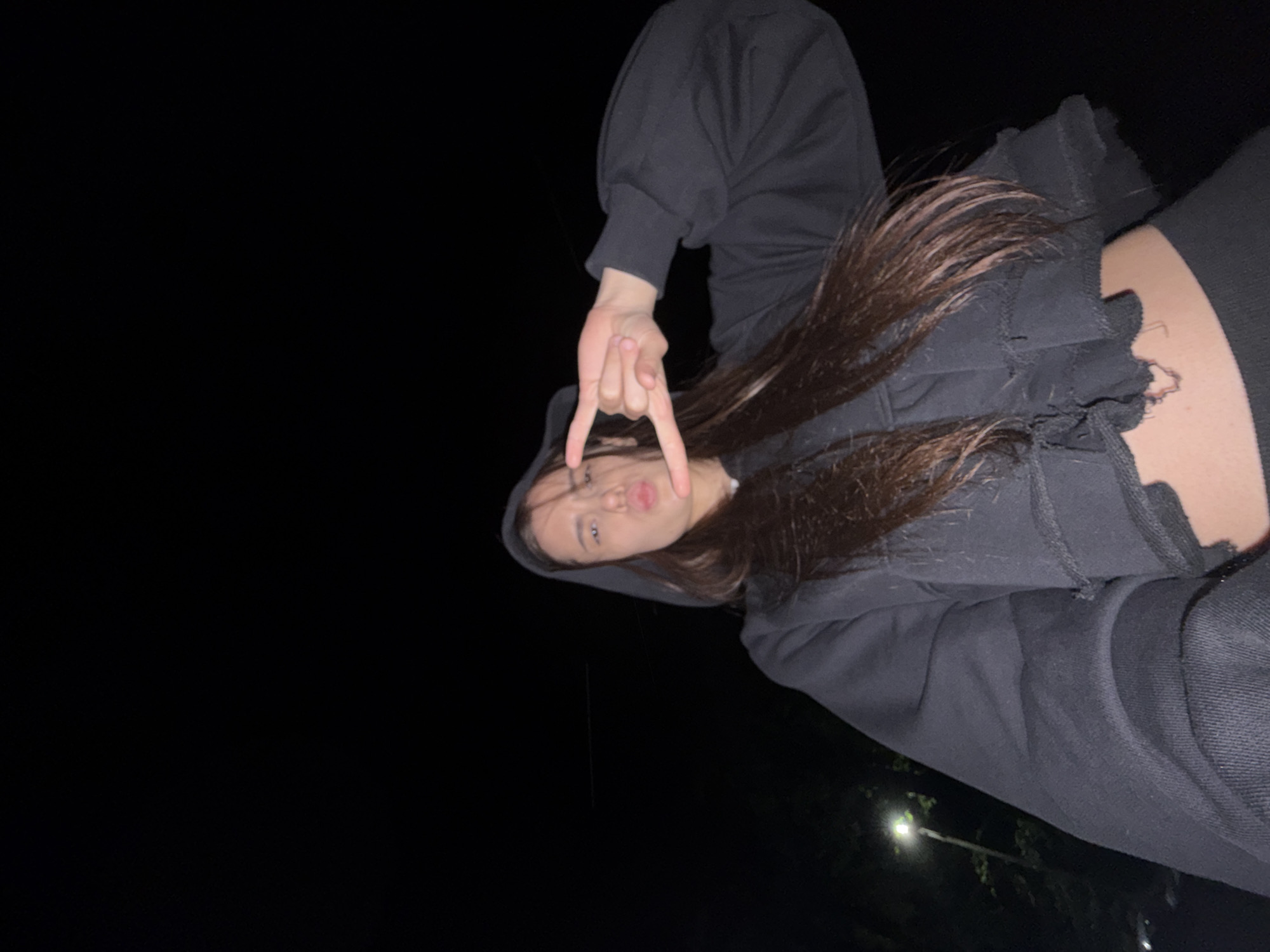 A woman wearing a dark hooded sweatshirt is pictured at night. She is making a hand gesture. The background is primarily dark, with a faint light source visible at the lower right. The photo was taken in Chaikovsky, Russia.