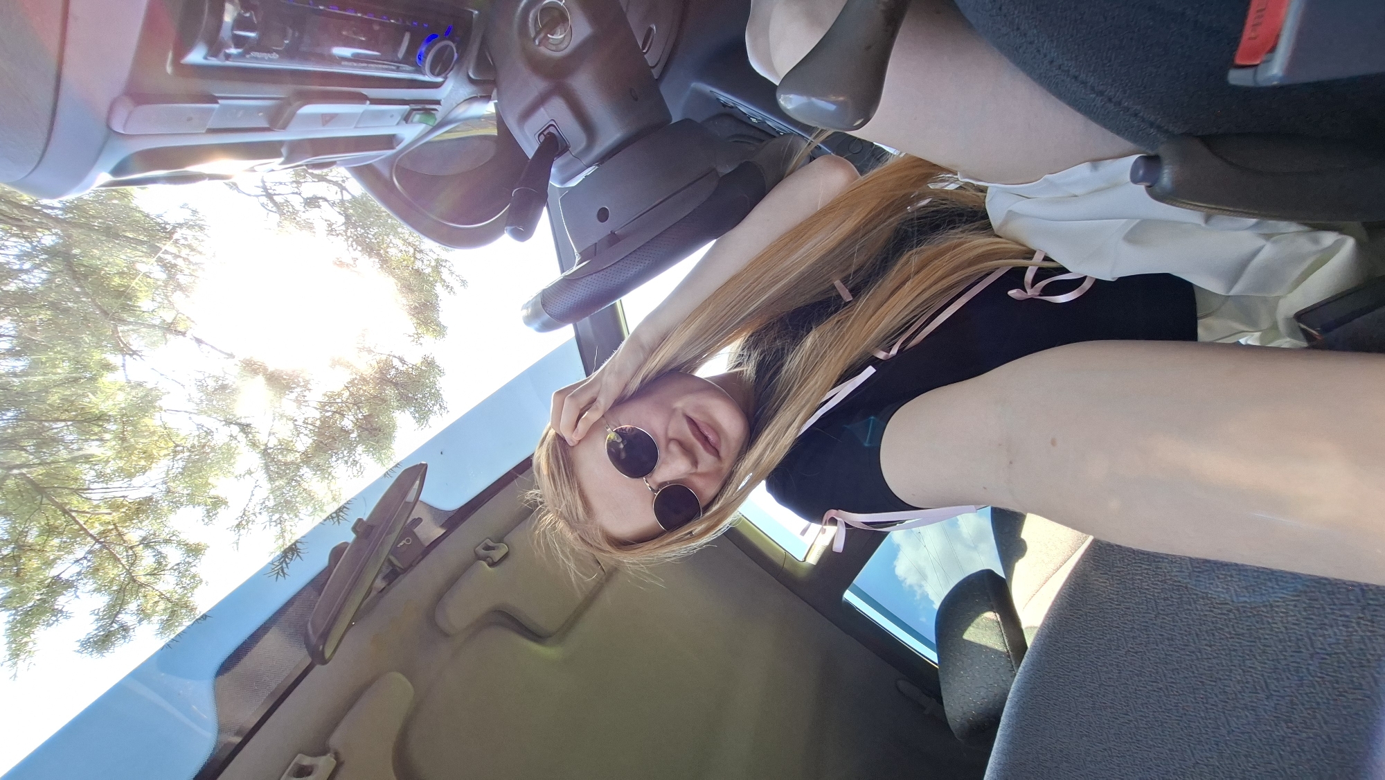 A young woman with long blonde hair and sunglasses is sitting in the driver's seat of a car, her hand resting on her head. The sun is shining brightly through the car's sunroof, casting a warm glow on the scene. The interior of the car is visible, including the steering wheel and dashboard. The woman is wearing a black t-shirt with pink ribbons. The background shows a glimpse of trees and a blue sky. The exact location in Bolshoy Bukor, Russia, cannot be determined from the image alone.