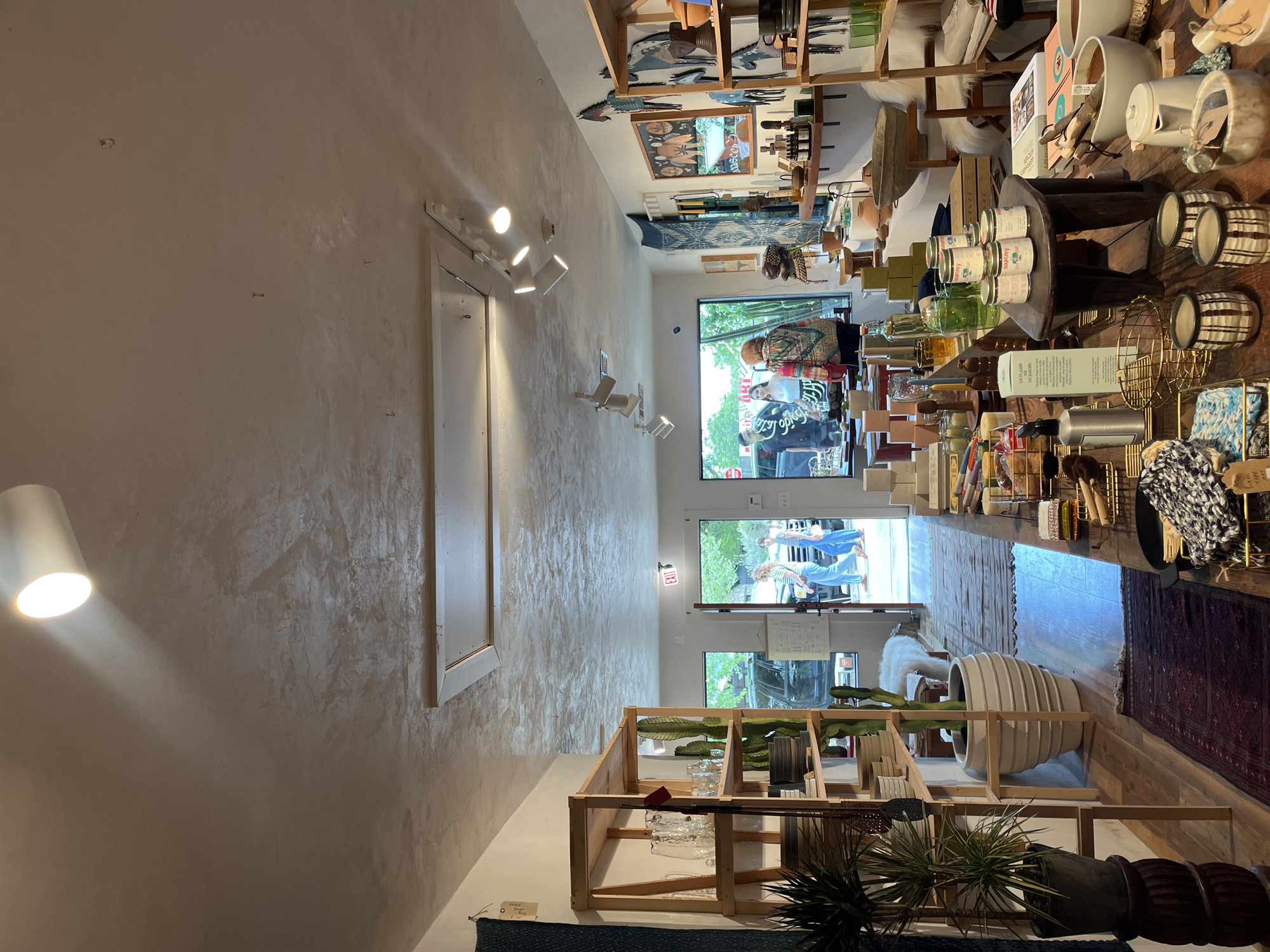 The image shows the interior of a retail store in Wimberley, Texas, United States. The space is narrow and long, with a white textured wall on the left and a wall of shelves stacked with merchandise on the right. At the back are windows and a doorway. People, including an older woman, are visible through the windows. There are various decorative objects on the shelves including pottery, plants, containers, and home goods. The lighting includes track lighting on the wall and recessed ceiling lights. An 'EXIT' sign is visible above the doorway.