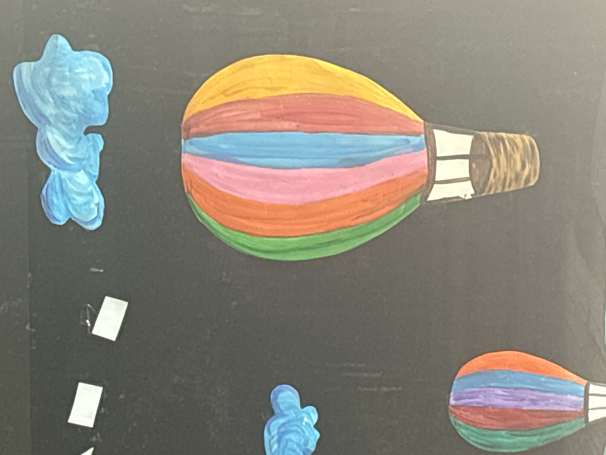 The image shows a portion of a dark background with several colorful, painted elements. In the center right, a large hot air balloon with vertical stripes of green, orange, pink, blue, red, and yellow dominates the frame. Below this balloon is a basket woven with brown and tan colors. To the lower left, a smaller hot air balloon with similar colorful stripes and a basket is partially visible. In the upper right, a blue, cloud-like shape is depicted. To the upper left, two small white rectangular shapes and a white triangular shape are visible, along with another smaller blue cloud-like shape to the left of the larger hot air balloon. The overall scene suggests a whimsical or artistic display, possibly a mural or a decorative piece. The image does not contain any information related to the city of Ob, Russia.