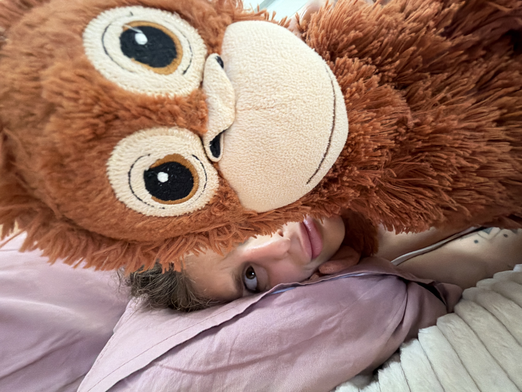 A young person with brown hair is lying in a bed with pink bedding in an indoor environment. A brown plush animal head is positioned over their face, partially obscuring it. The exact location is North Kuta, Indonesia.