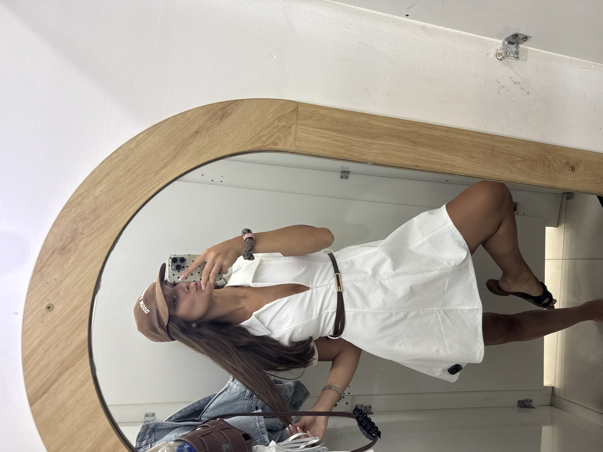 A female is taking a selfie in front of an oval mirror with a light wood frame. She's wearing a brown baseball cap, a white belted dress, and sandals. She is holding a smartphone with a multicolored case up to take the photo. Reflected below her in the mirror are various items, including a denim jacket, a brown bag, a white ribbon or tag, and a black hair tie. The environment appears to be an indoor space, possibly a fitting room or closet, with white walls. The photograph's background suggests it might be taken in a city, but not enough information to identify Da Nang specifically in Vietnam.