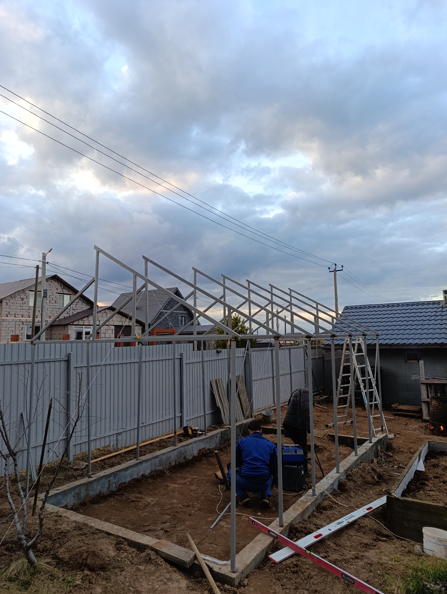 This outdoor scene captures a construction site, likely a backyard or garden, in Chaikovsky, Russia, under an overcast sky. The primary activity involves two individuals erecting a metal framework, possibly for a greenhouse or shed.
One person, dressed in a blue jacket and blue pants, is crouched on the dirt ground, seemingly working with a welding machine or similar electrical equipment positioned before them. A second person, partially obscured, stands nearby in a dark jacket. The metal structure, comprising vertical posts and angled roof beams, is partially assembled, extending across the image from left to right. A prominent red and white spirit level, with "Empire" faintly visible on its white section, lies diagonally in the immediate foreground. A silver metal ladder is propped against the partially built frame on the right.
The ground is disturbed earth, indicative of ongoing work, with scattered construction materials such as wooden planks and metal sheets. A corrugated metal fence forms a barrier across the mid-ground. Beyond the fence, several houses are visible, one appearing to be under construction with exposed block walls, and another featuring a dark tiled roof. Utility poles and overhead power lines crisscross the cloudy sky. To the far right, a small fire burns within a metal drum or pit. Bare tree branches are visible in the left foreground. The sky is filled with grey, patchy clouds, creating soft, diffused lighting.