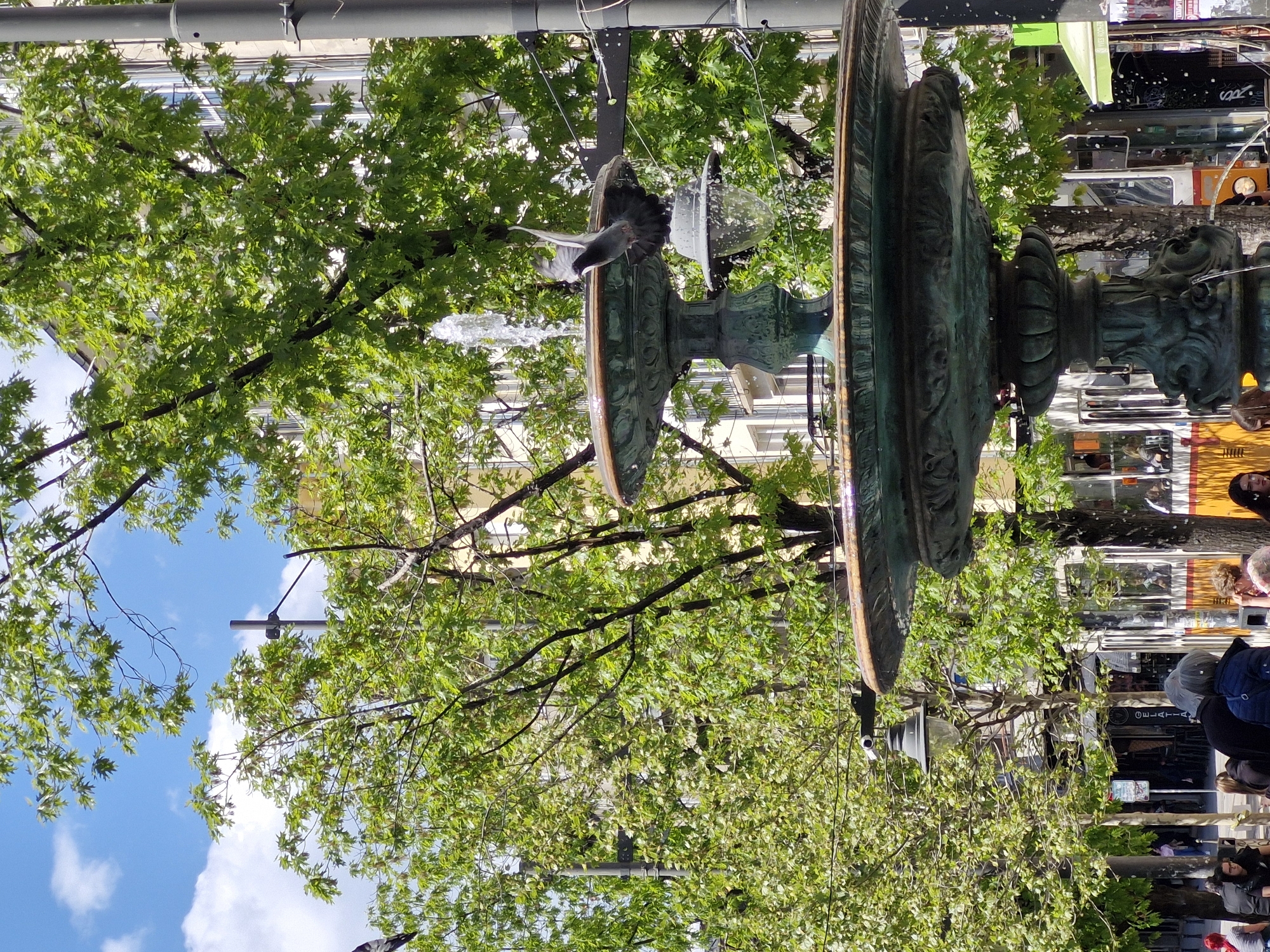 The image depicts a fountain in the city of Sofia, Bulgaria. The fountain is ornate and made of dark metal, with multiple tiers and intricate carvings. Water is flowing from the fountain. A bird is captured mid-flight near the water. The fountain is surrounded by trees with green foliage, and the sky is visible with some clouds. In the background, there are buildings and people walking by, suggesting a busy urban environment. Some people are standing around the fountain. There is a bus in the background. A street lamp is present next to the fountain.