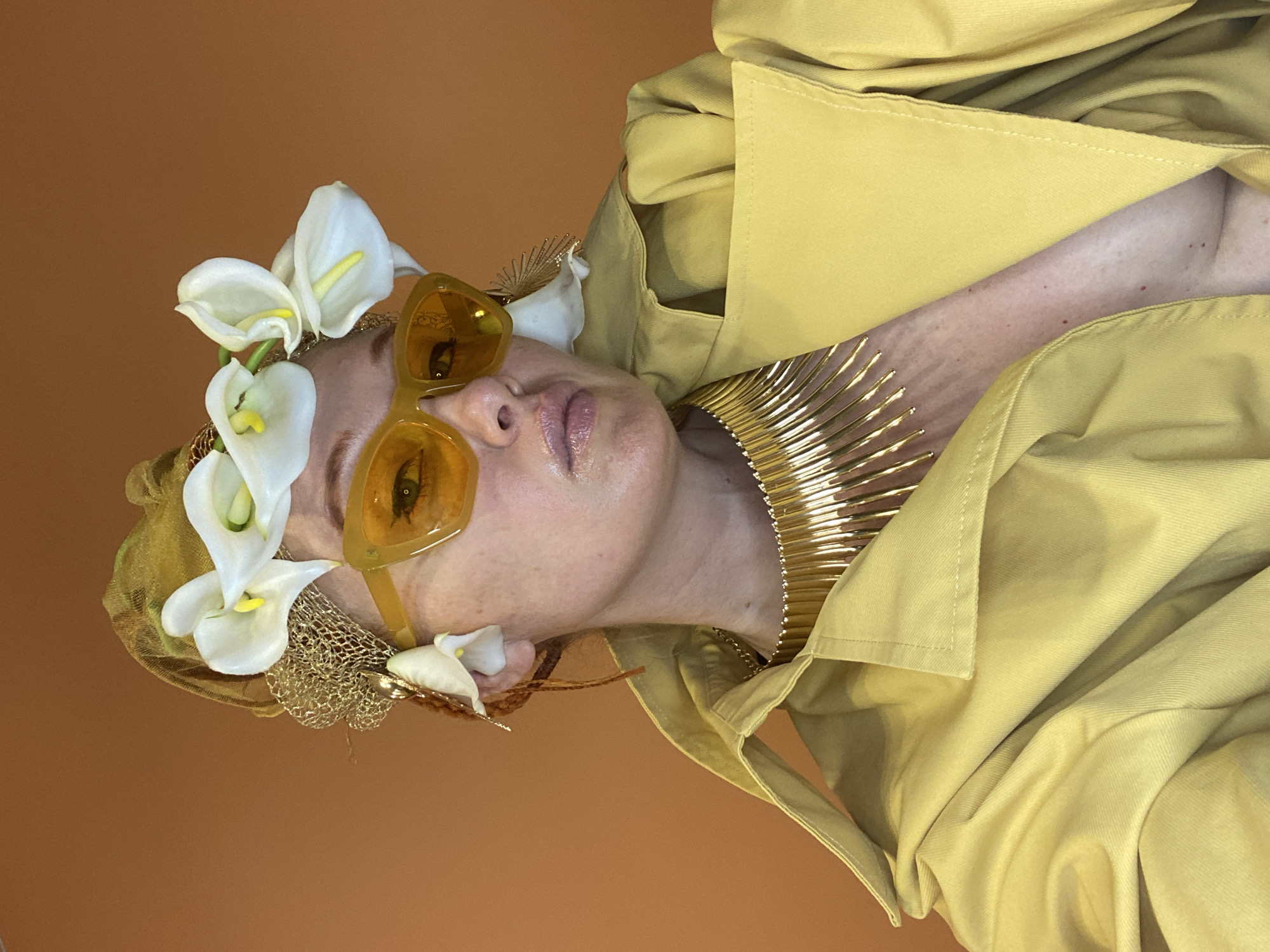 This is a studio portrait of a person with their head tilted to the side, against a plain brown background. The subject wears a yellow trench coat, large orange-tinted sunglasses, and a gold necklace. They are adorned with white calla lilies and a gold mesh headpiece, suggesting a stylized or artistic presentation. The lighting is bright and even, typical of a controlled studio environment. The subject's facial expression is neutral, and their makeup appears subtle with a hint of lip gloss. The overall aesthetic is avant-garde and fashion-forward, with a focus on bold accessories and a monochromatic color scheme of yellow and gold. No text is visible in the image.