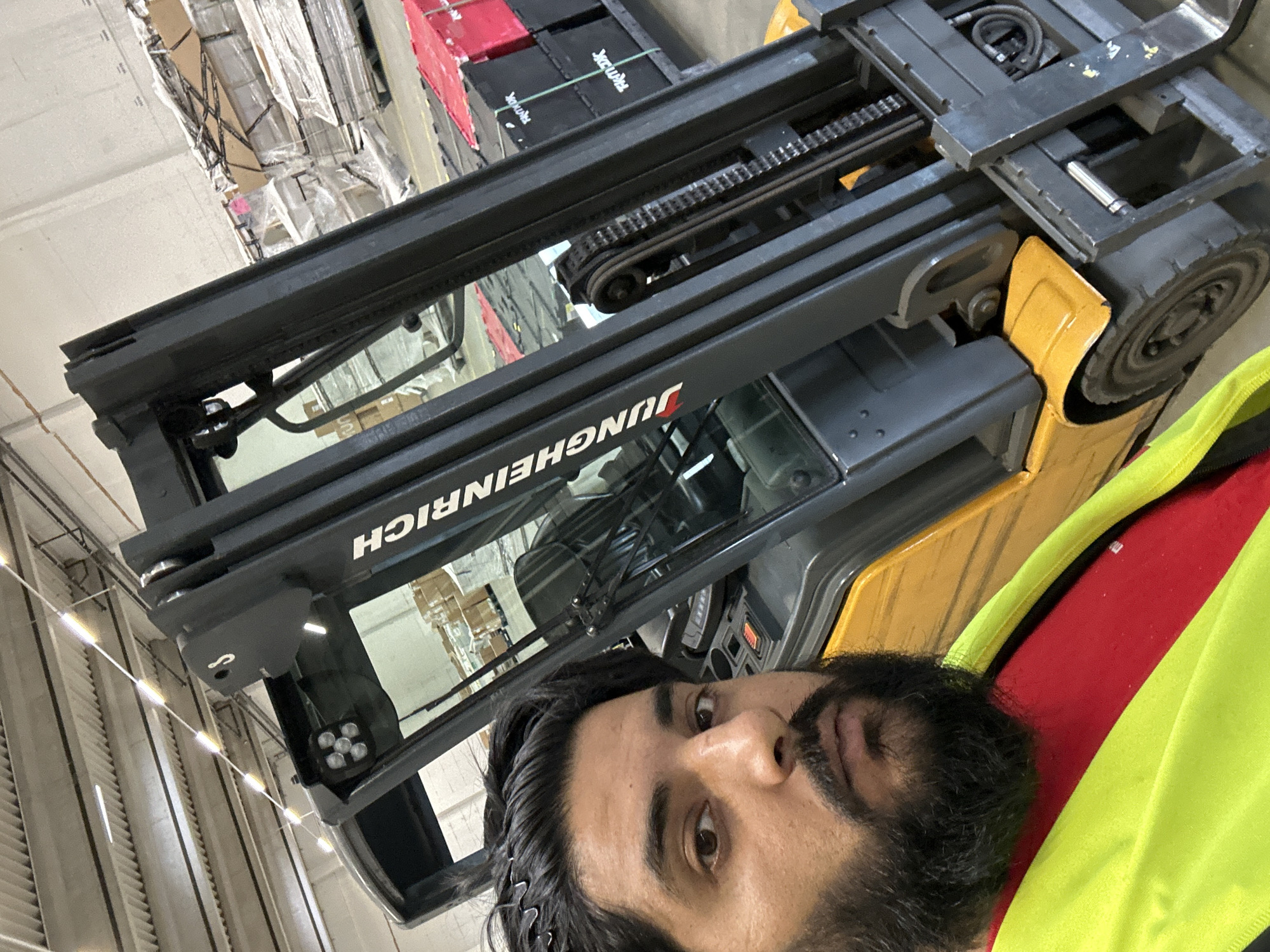 A man with dark hair and a beard, wearing a red shirt and a yellow high-visibility vest, is taking a selfie in front of a yellow and black forklift. The forklift has the brand name "Jungheinrich" visible on its mast. The background appears to be a warehouse or industrial setting, with shelves stacked with boxes and pallets of goods. Red plastic containers are also visible. The ceiling has visible light fixtures and structural elements. The scene is likely in Schweitenkirchen, Germany, as indicated by the user prompt, implying a commercial or industrial location within that area.