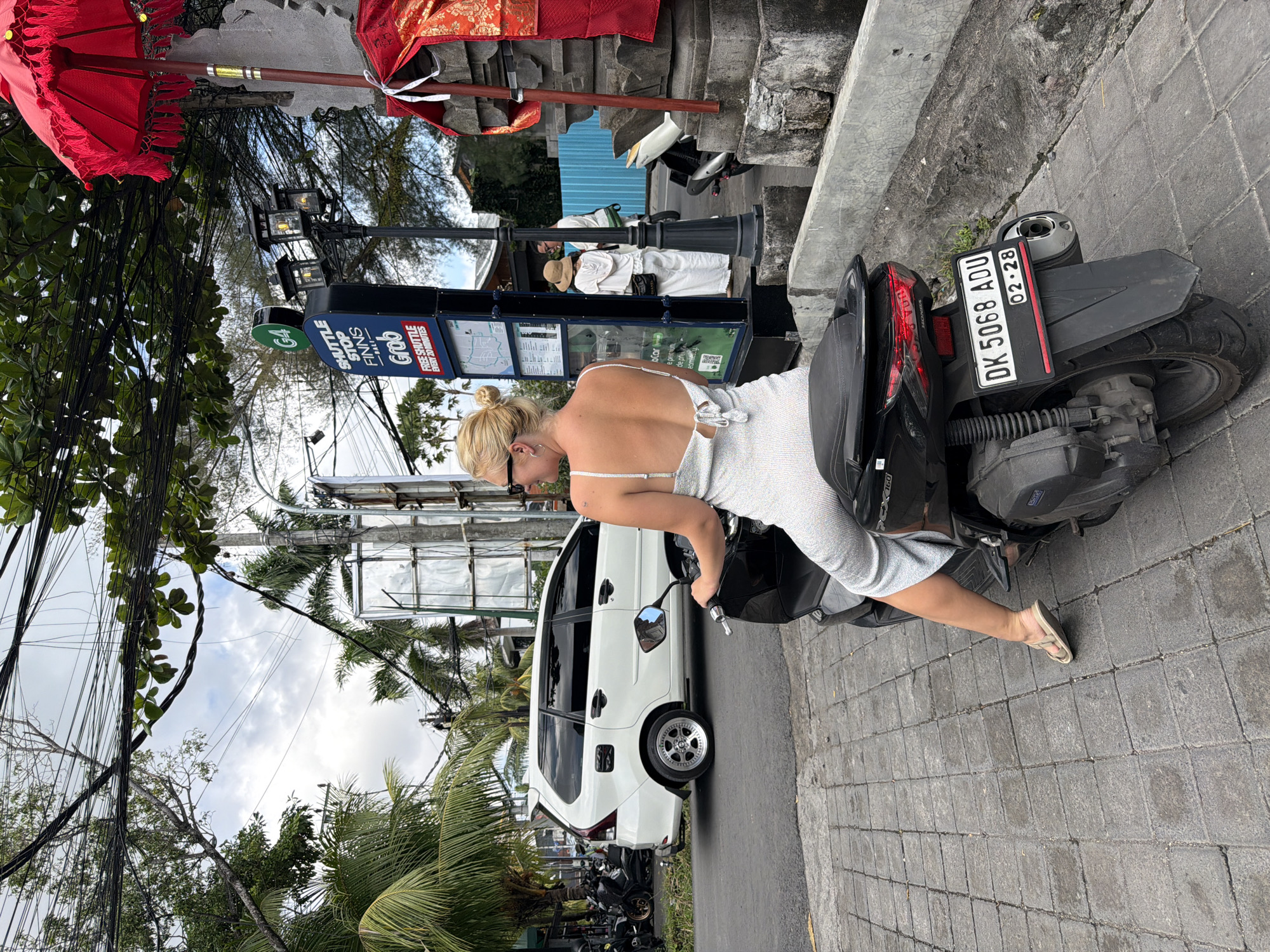 A woman with blonde hair, wearing a sparkling white dress and sandals, is sitting on a black Honda PCX160 scooter. The scooter's license plate reads "DK 5068 ADU 02-28." The scene appears to be in North Kuta, Indonesia, based on the "FINNS BALI" sign. A white van is parked next to the scooter, and several other scooters are parked nearby. There's a "SHUTTLE STOP" sign for a "Grab" shuttle, and another structure with red fabrics and a red umbrella. Two individuals dressed in white attire are standing near the shuttle stop.
