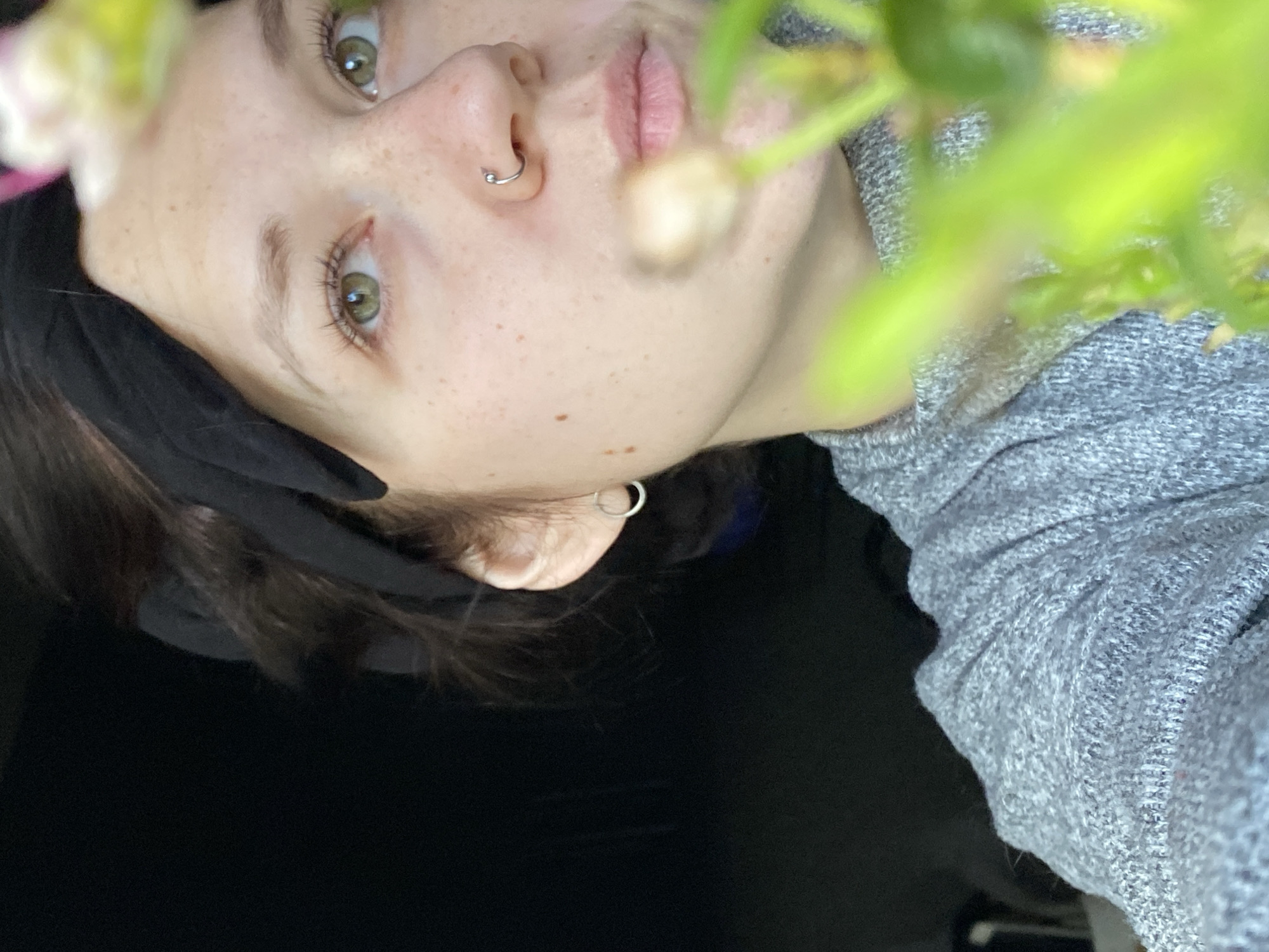 A close-up, low-angle, and slightly tilted portrait shot depicts a young woman with fair skin and green eyes. She has freckles on her face and a nose ring. She is wearing a gray knit sweater and a black headband. She also has a hoop earring. The background is dark, and part of a flowering plant is visible in the foreground. No evidence suggests this image was taken in the city of Chaikovsky, Russia.