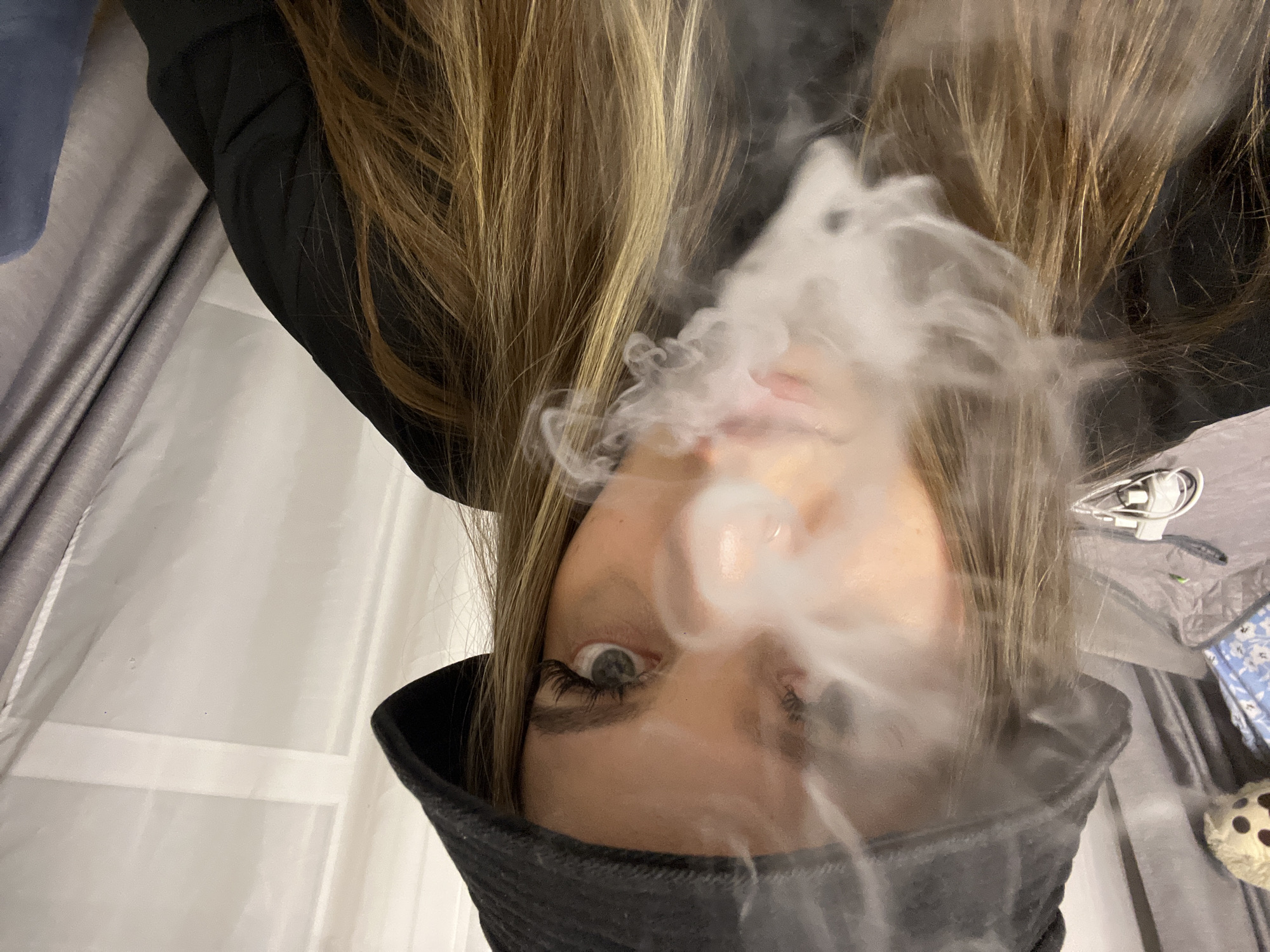 The image, taken in Krasnodar, Russia, is rotated 90 degrees counter-clockwise, presenting a close-up of a young person exhaling a dense plume of white smoke or vapor. The smoke, forming a ring-like shape, originates from their mouth, obscuring much of their nose and lips, suggesting the use of a vape device or similar.
The subject is primarily seen from the chin upwards, wearing a dark top, possibly a hooded jacket, with a collar-like feature pulled up to their lower face. Long, light brown hair cascades towards the top of the frame. One eye, adorned with prominent eyelashes, is clearly visible and appears focused on the act of exhaling.
The setting is indoors, with various domestic elements in the background. To the left, there are translucent light-colored curtains or blinds. Grey fabric, resembling bedding or a blanket, is visible on both sides of the subject. In the upper right background, white electrical cords and a charger are present. A light brown object with dark polka dots, possibly a patterned pillow or plush toy, is situated in the lower right corner. The indoor lighting suggests the image was taken in the evening or at night. No discernible text is visible in the image.