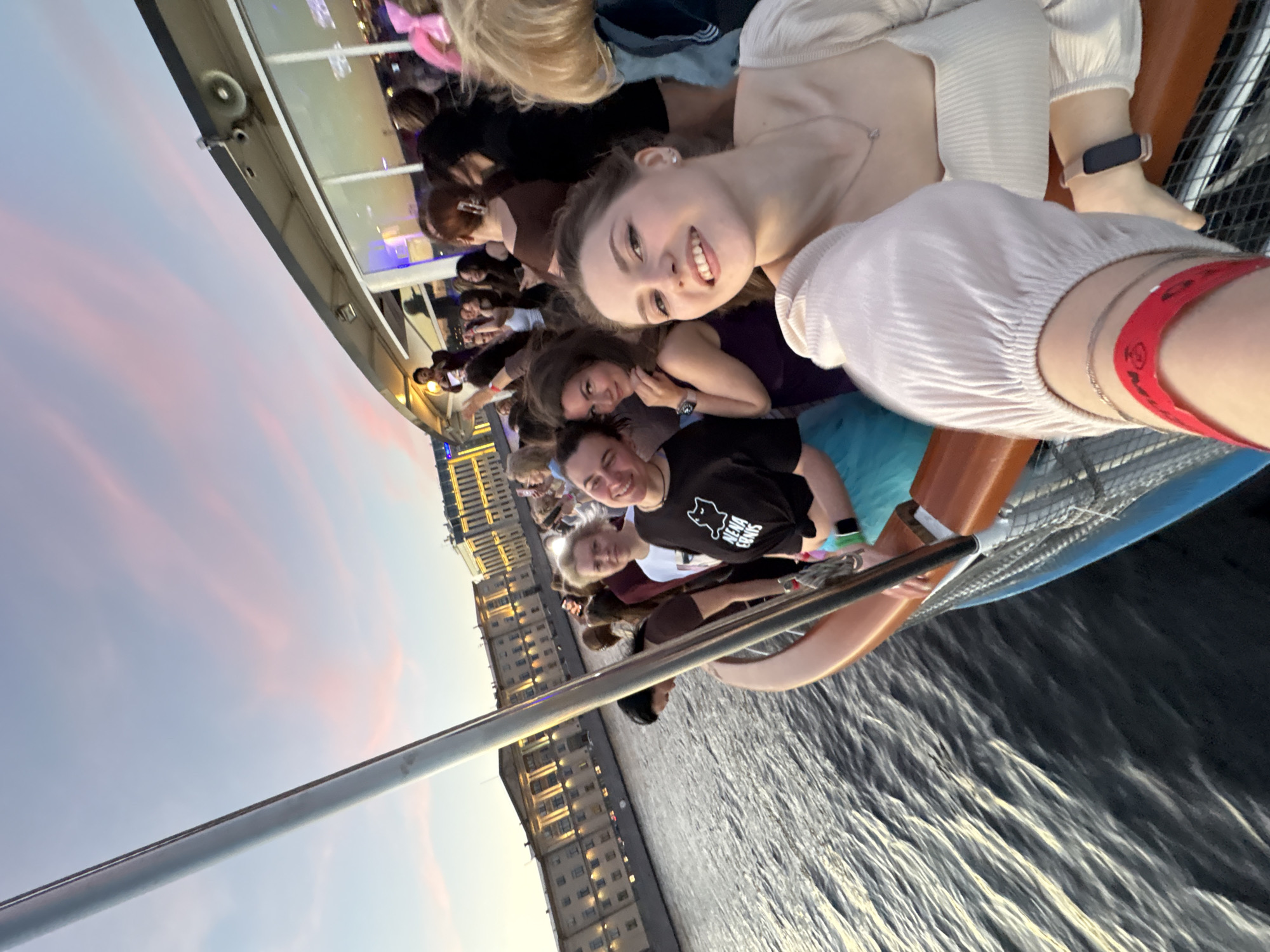 The image depicts a group of people on a boat sailing on a river in Saint Petersburg, Russia. The boat is traveling along the Neva River, with the buildings of the city visible in the background. The sky is illuminated with the soft hues of sunset, casting a warm glow over the scene. The people on the boat appear to be enjoying themselves, with some posing for photos and others conversing. One woman in the foreground is taking a selfie, smiling brightly. The overall atmosphere is festive and relaxed, suggesting a pleasant outing on the water. The architecture visible in the background is characteristic of Saint Petersburg, with its grand imperial buildings.