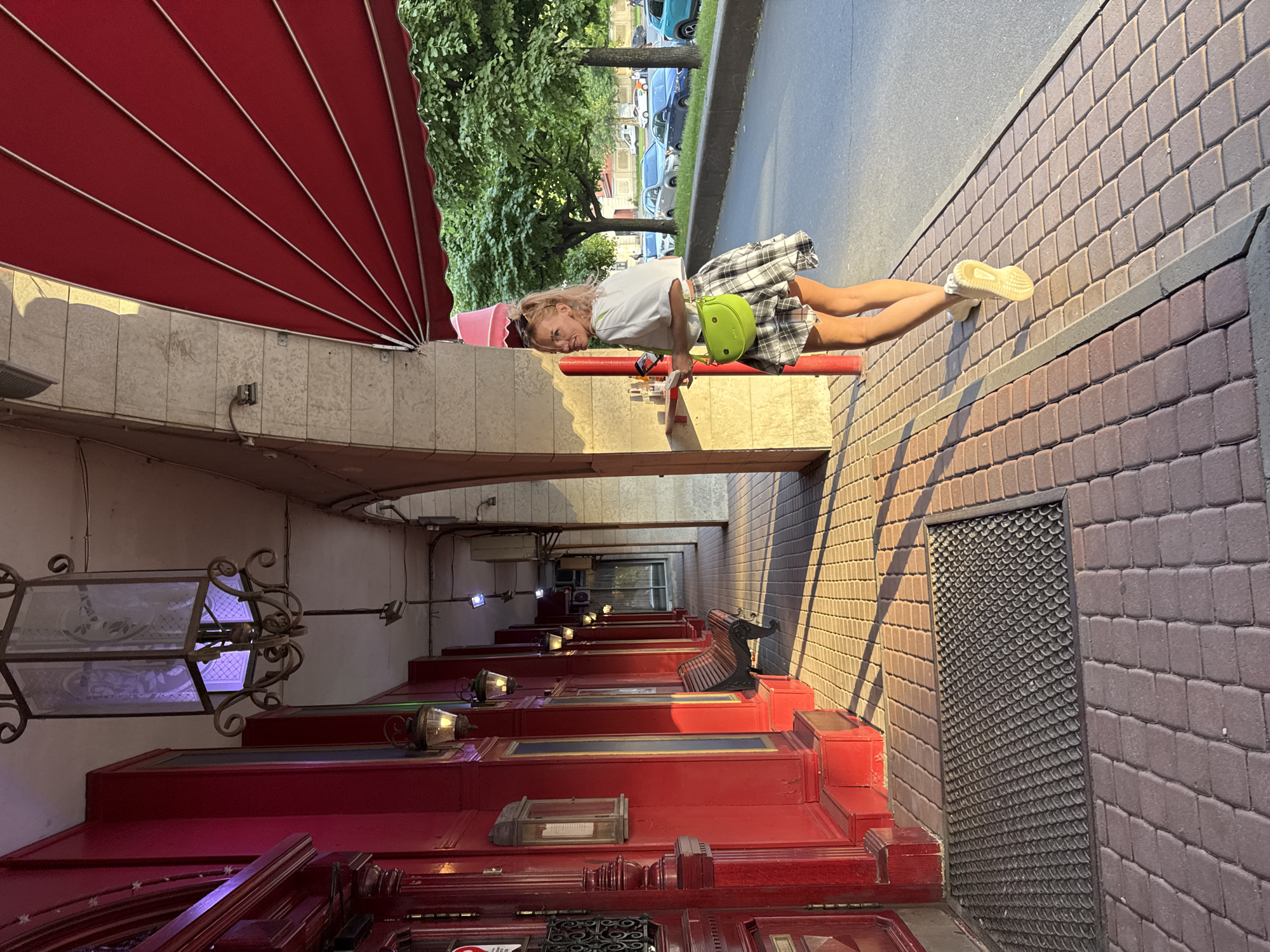 A blonde woman with a green purse and a plaid skirt walks on a sidewalk in Saint Petersburg, Russia. The woman is wearing a white crop top and yellow sneakers. She is walking away from the camera, but looking over her left shoulder. The sidewalk is made of red brick pavers, and there is a red mat in front of a building with red walls. An awning is above the woman. Trees and cars are visible in the background. The sun is casting long shadows on the sidewalk.