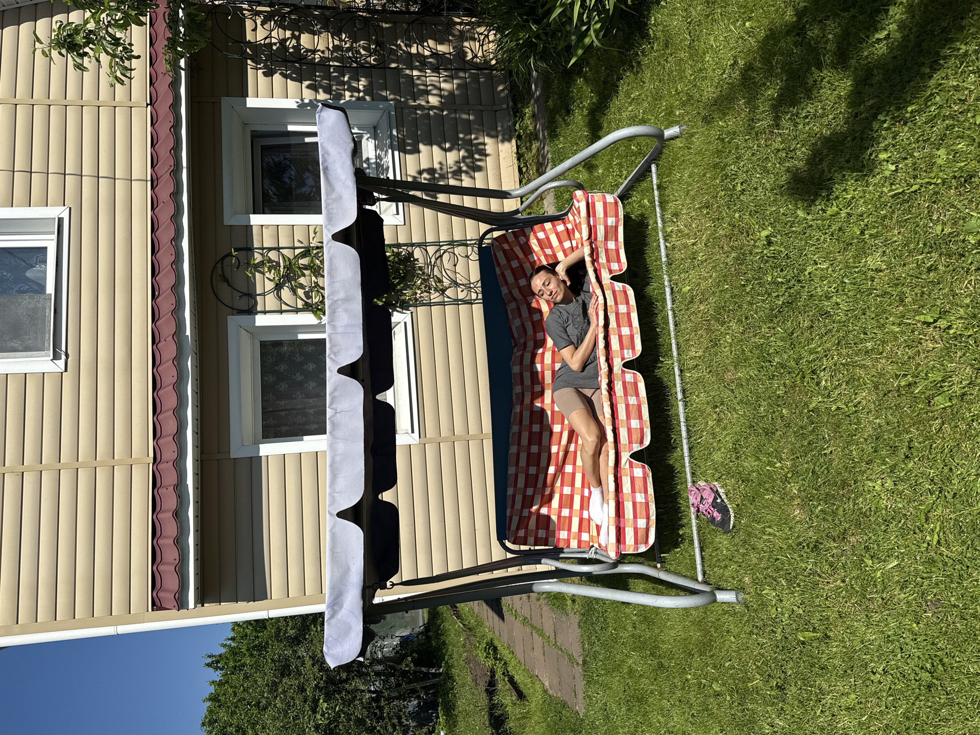 This image depicts a sunny outdoor scene in a residential or suburban garden setting. A female, appearing to be an adult, is resting on a garden swing. She is lying on her right side on the swing's red and white checkered patterned mattress, wearing a grey t-shirt, grey shorts, and white socks. Her eyes are closed, and her right hand is positioned under her head, suggesting she is sleeping or relaxing.
The garden swing features a silver-colored metal frame and a grey fabric canopy overhead. Directly beneath the front-left leg of the swing, a pair of dark athletic shoes with pink accents is placed on the grass.
In the background, a house, possibly two stories high, is visible. Its exterior is clad in light beige horizontal siding, and it has a reddish-brown corrugated metal roof. Multiple white-framed windows are present on the house wall. Ornate dark metal trellises are attached to the house facade, with some green foliage growing on at least one of them.
The ground is covered with vibrant green grass, indicating a well-maintained lawn. To the far left, a path constructed from stone or concrete pavers is partially visible. Trees and additional greenery are present in the background and surrounding areas. The scene is brightly illuminated by direct sunlight, casting distinct shadows from the swing and other elements onto the grass and the house, indicating a clear sky. While the prompt specifies the location as Saint Petersburg, Russia, the visual information within the image itself does not contain specific identifying landmarks or features to confirm this precise geographical location; the setting is typical of a dacha or suburban private garden.