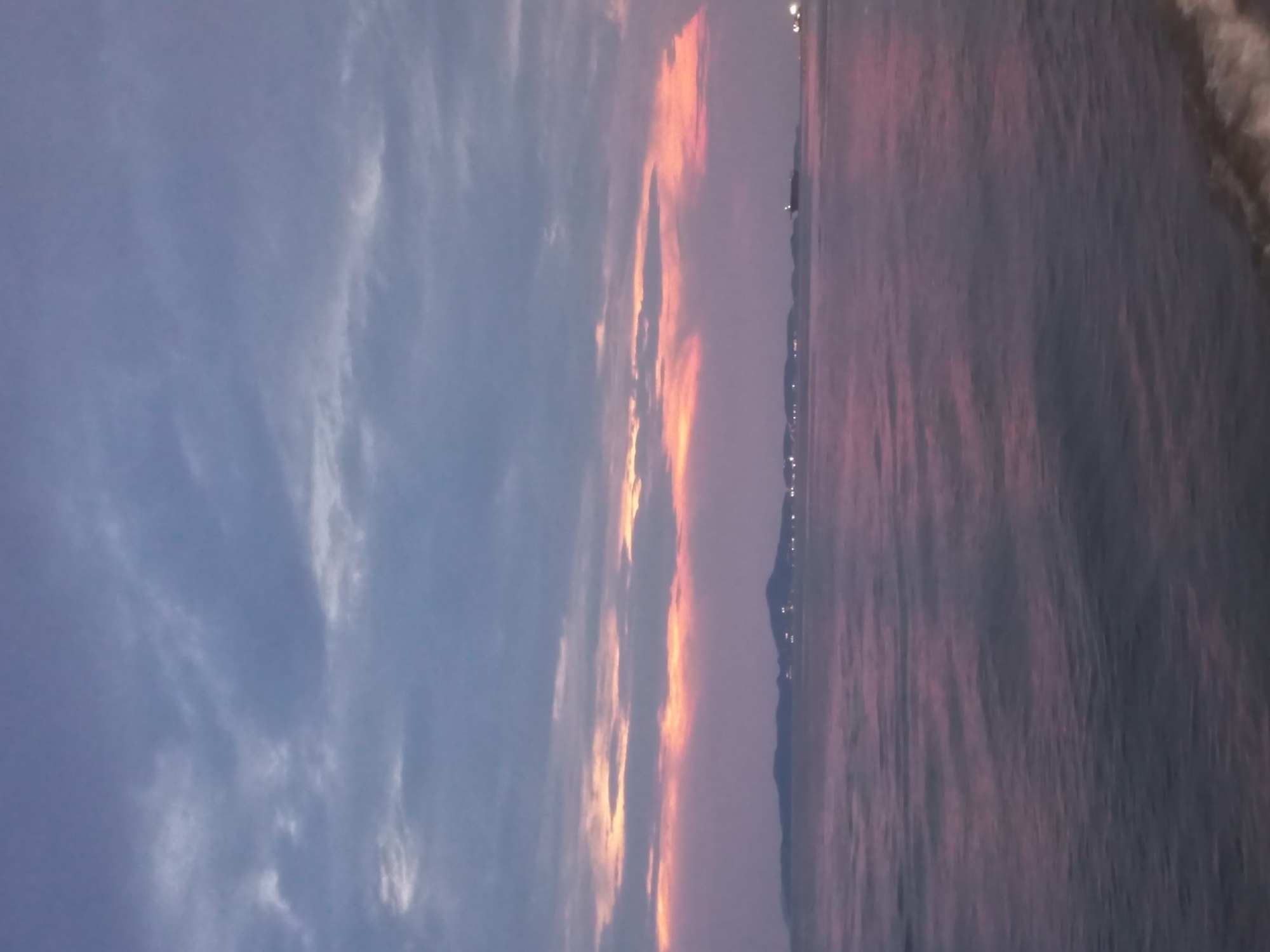 The image depicts a sunset scene in Pattaya City, Thailand. The sky is filled with clouds that transition from a muted blue-gray on the left to fiery orange and pink hues in the center, suggesting the presence of the setting sun. The sea occupies the right side of the frame, reflecting the colors of the sky and creating a rippled effect. Along the horizon, there are faint lights indicating the city and possible structures or ships. The water's edge is visible in the upper right, where waves gently break on the shore.