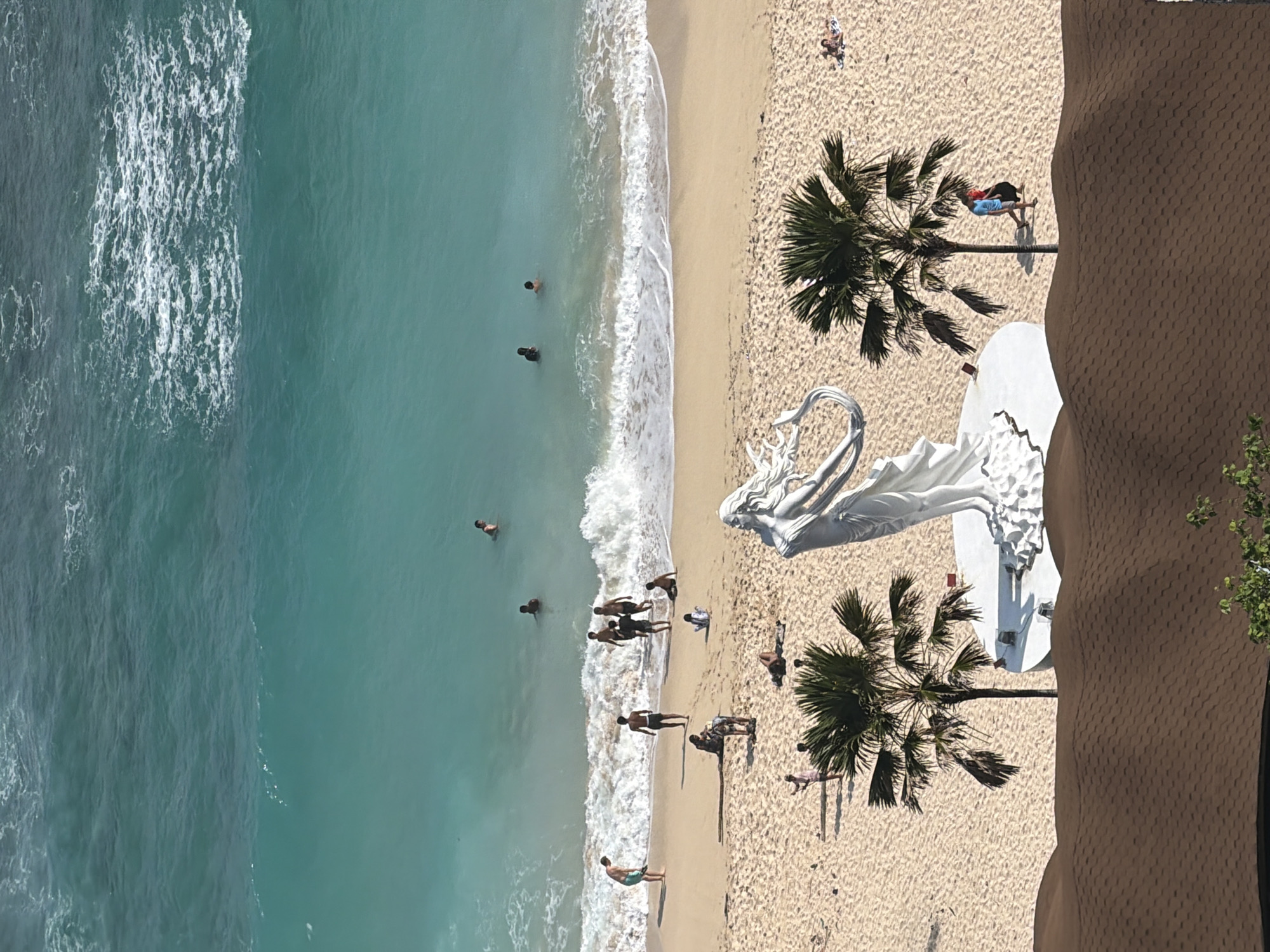 An elevated, high-angle photographic image displays a coastal environment in Ungasan, Indonesia. The upper portion of the scene features a body of turquoise ocean water, exhibiting varying shades of blue and green, with white foamy waves breaking at the shoreline. Six individuals are discernible within the ocean, either wading or swimming.
The middle ground is occupied by a wide expanse of light tan sand. On this beach, approximately twenty individuals are visible. Five persons are positioned at the water's edge, interacting with the incoming waves. Further inland on the sand, two distinct palm trees frame a central, large white statue of a draped female figure standing on a circular white base. Near the base of the right palm tree, two individuals walk toward the viewer. Another individual lies prone on a towel near the right edge of the frame. Scattered other individuals are observed sitting or standing across the sandy area.
The bottom foreground of the image partially shows a reddish-brown roof structure characterized by undulating, shingle-like textures. Minimal green foliage is visible at the extreme bottom edge. The scene is illuminated by direct sunlight from a high angle.