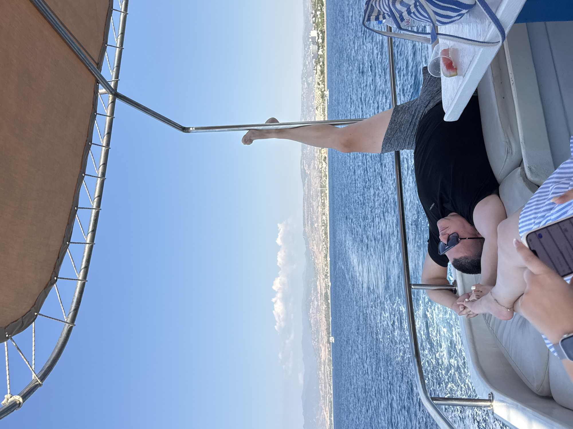 A man and woman are relaxing on a boat with the coastline of Neo Chorio, Cyprus visible in the background. The man is lying down, wearing a black t-shirt and sunglasses, with one leg raised in the air. The woman's feet are visible in the foreground, and she appears to be holding a phone. A table beside them holds a glass with a slice of watermelon and a striped bag. The boat is on the blue sea, under a clear sky.