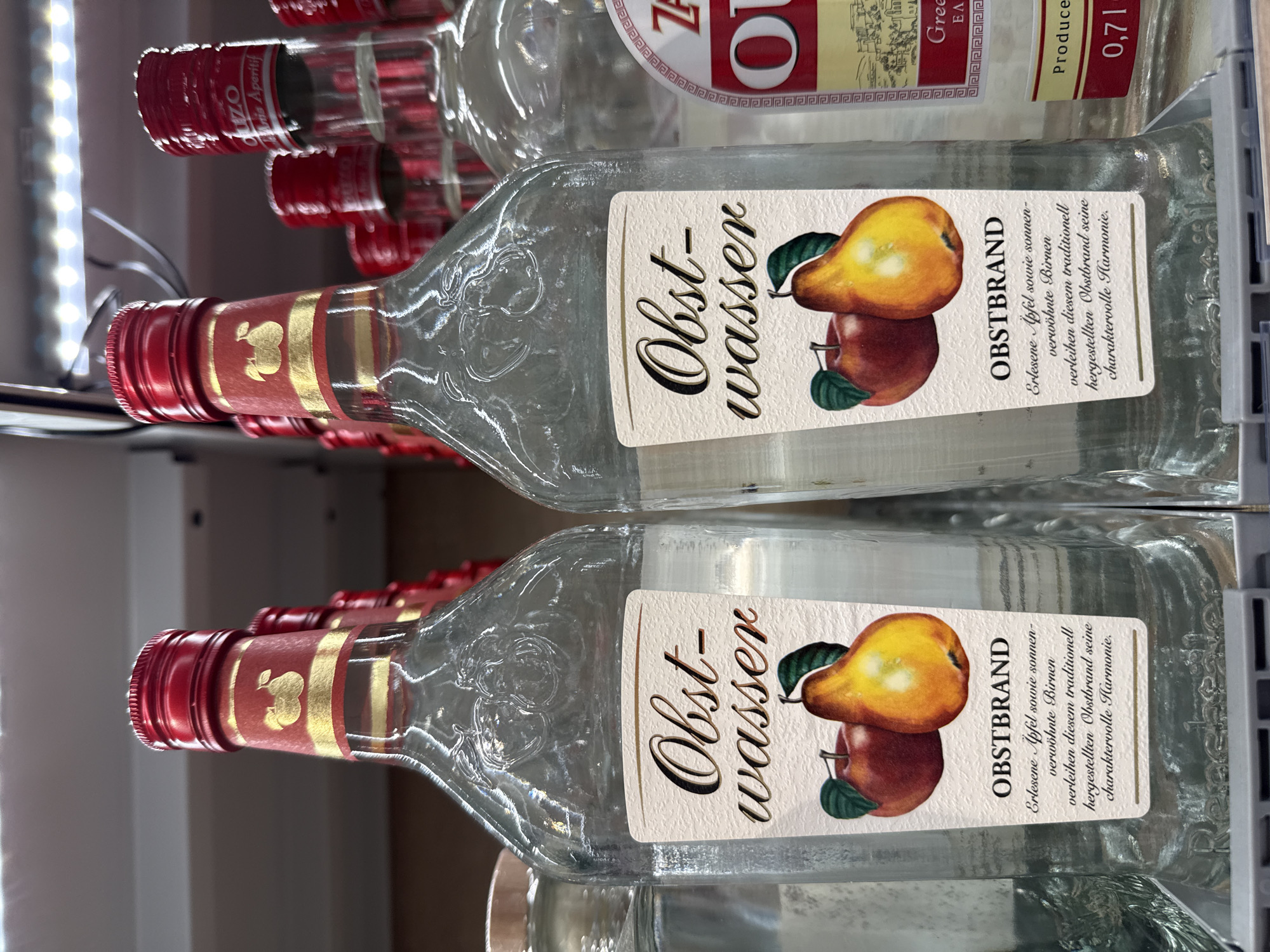 Two clear glass bottles of "Obstwasser" (fruit water) are prominently displayed side-by-side on a shelf in a retail setting, likely a liquor store or supermarket. The bottles have red caps and gold labels featuring an illustration of an apple and a pear. The labels also contain German text describing the product as "Obstbrand" (fruit brandy) made from selected apples and sun-ripened pears. The bottles are filled with a clear liquid. To the right of the "Obstwasser" bottles, parts of other bottles, including one with a label indicating "Ouzo", are visible. The background shows additional bottles on shelves and overhead fluorescent lighting. The setting appears to be indoors in a commercial establishment. The specific location in Bad Lauterberg im Harz, Germany, is not discernible from the image itself.