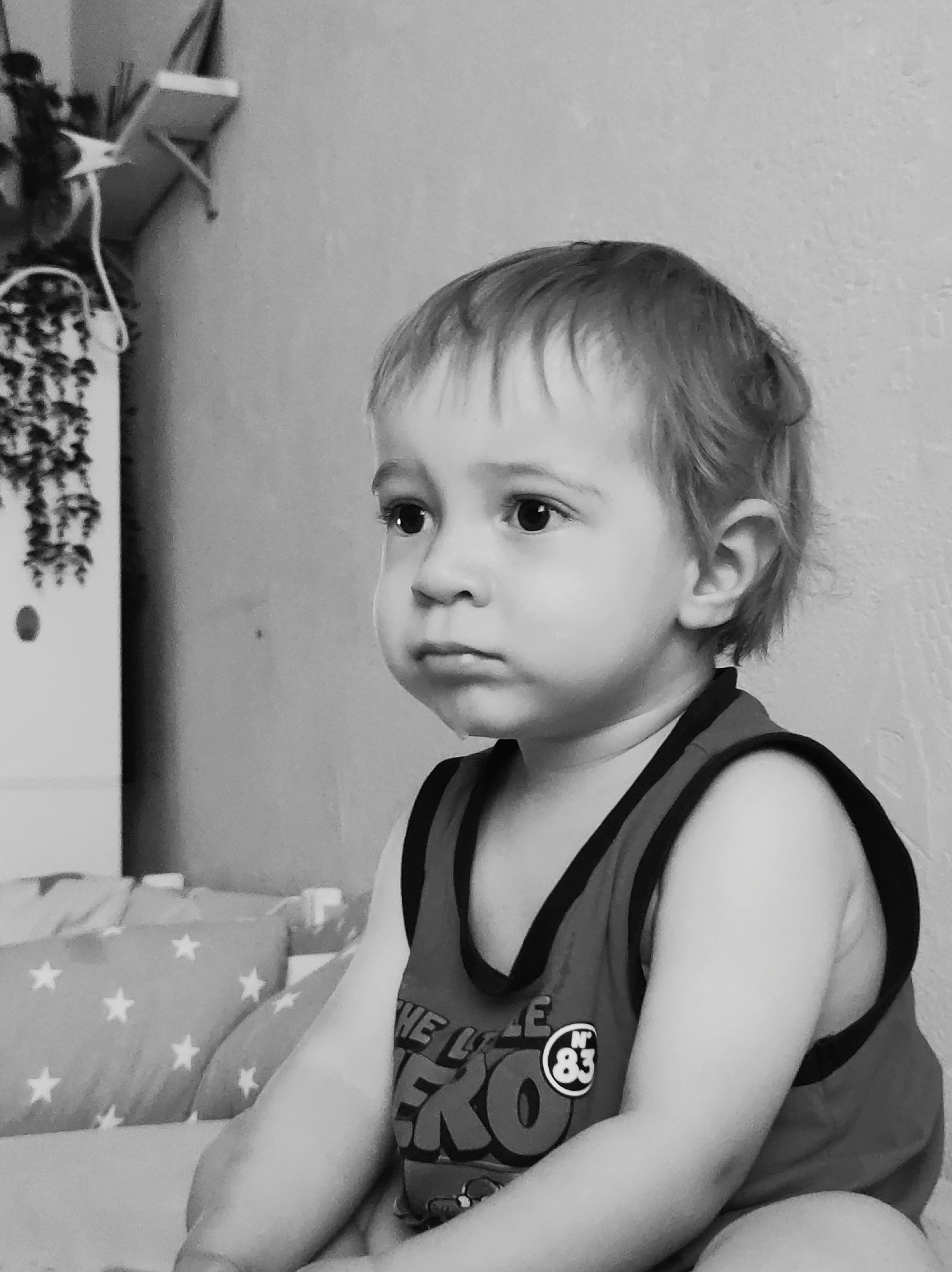 A monochrome, medium shot depicts a toddler seated indoors. The child is wearing a sleeveless top with a dark trim and a printed design featuring the word "HERO" and the number "83" inside a circular badge. The toddler has a somber expression, with slightly pursed lips and eyes gazing towards the left of the frame. The background includes a wall, bedding, and pillows patterned with stars. To the left, a shelf-like structure with hanging decorations is visible. There is no specific evidence to confirm the image was taken in Chaikovsky, Russia.
