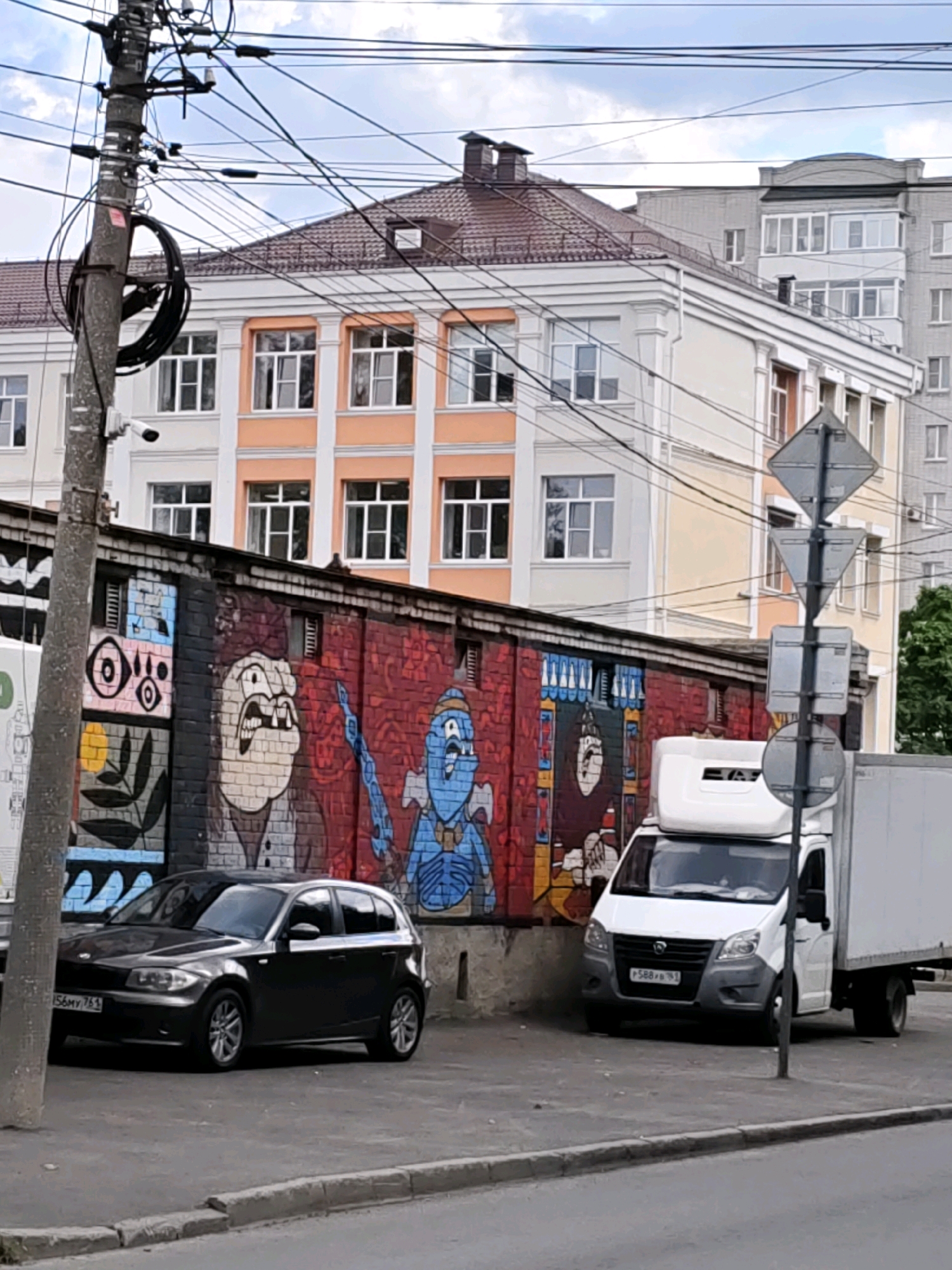 The image depicts an urban scene with a street, a brick wall covered in graffiti, and buildings in the background. A dark-colored hatchback car is parked on the left side of the street, adjacent to the graffiti wall. To the right of the car, a white delivery truck is parked. The graffiti on the wall features various cartoonish figures and abstract designs. The background includes a multi-story building with a light-colored facade and orange accents, and a taller, light gray apartment building. Utility poles with numerous wires are visible in the foreground, obscuring parts of the sky and buildings. The scene appears to be captured during the daytime under an overcast sky. The license plate on the car is partially visible and appears to be in Cyrillic script. The license plate on the truck is also visible and in Cyrillic script. The location is identified as Rostov-on-Don, Russia.