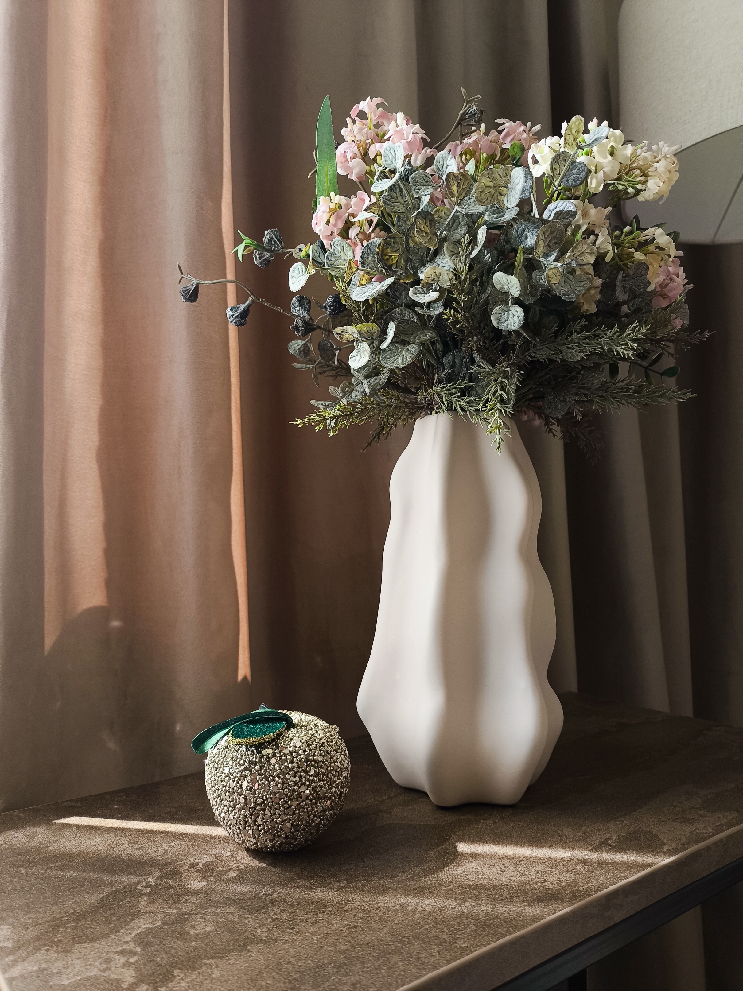 An indoor still life scene displays a decorative arrangement on a table against a backdrop of draped fabric. On the right, a tall, irregular-shaped vase holds a bouquet of artificial flowers and greenery. The flowers appear to be a mix of pale pink, white, and cream, interspersed with grey-green foliage and dark, twig-like accents. To the left of the vase, a small, spherical decorative object, possibly an apple, is covered in reflective, silvery beads and adorned with a green ribbon. The table surface is a textured, dark brown material. Sunlight casts a distinct shadow and highlights across the scene, indicating natural light entering from the left. The overall lighting is soft, with shadows defining the forms of the objects. The background consists of layered drapes in muted brown and pinkish-brown tones.