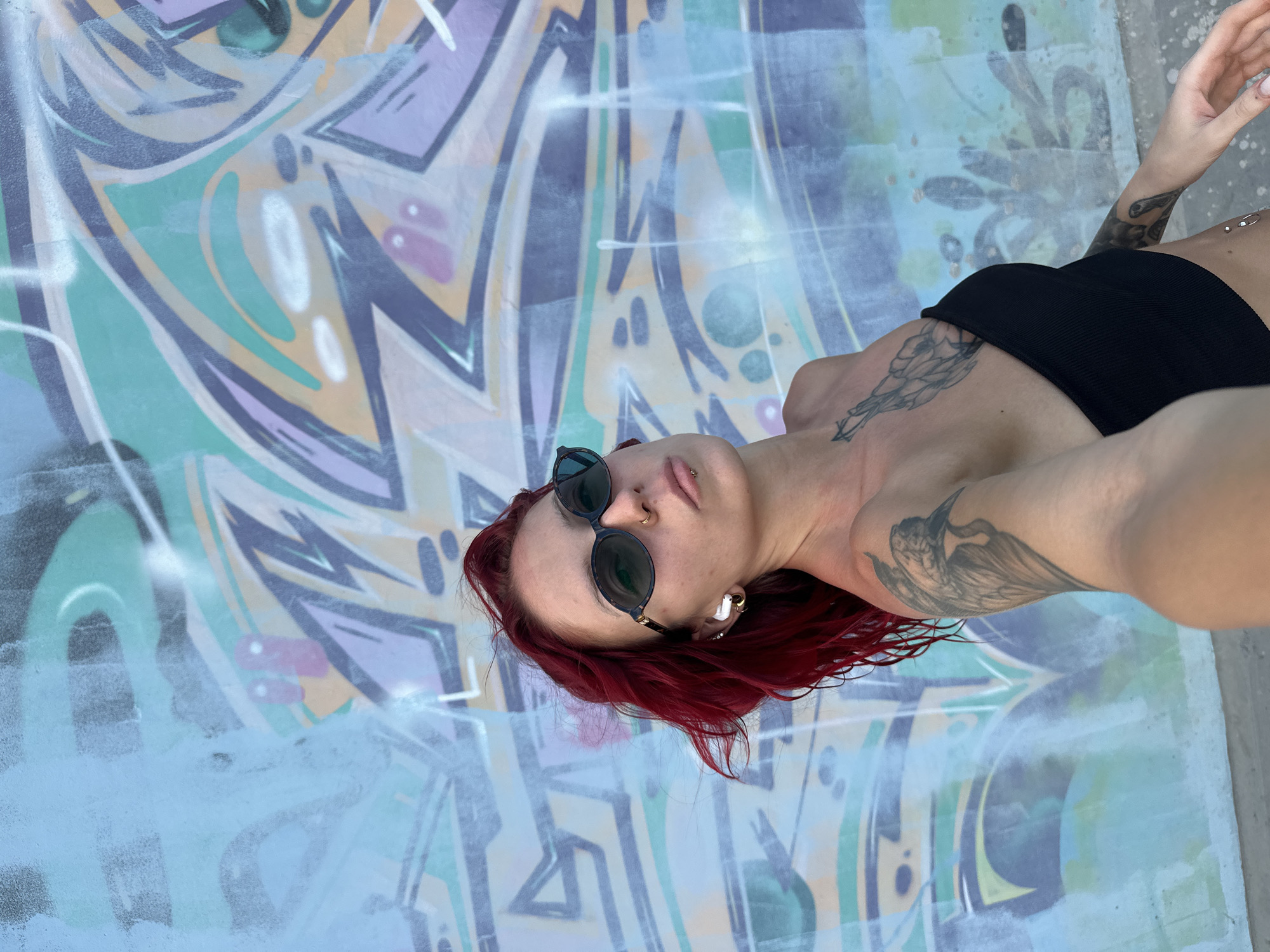 A woman with red hair and sunglasses, wearing a black top, is standing in front of a graffiti wall in Tibubeneng, Indonesia. She has multiple tattoos on her arms and chest, as well as piercings in her nose and ear. The graffiti wall features a variety of colors and abstract shapes.