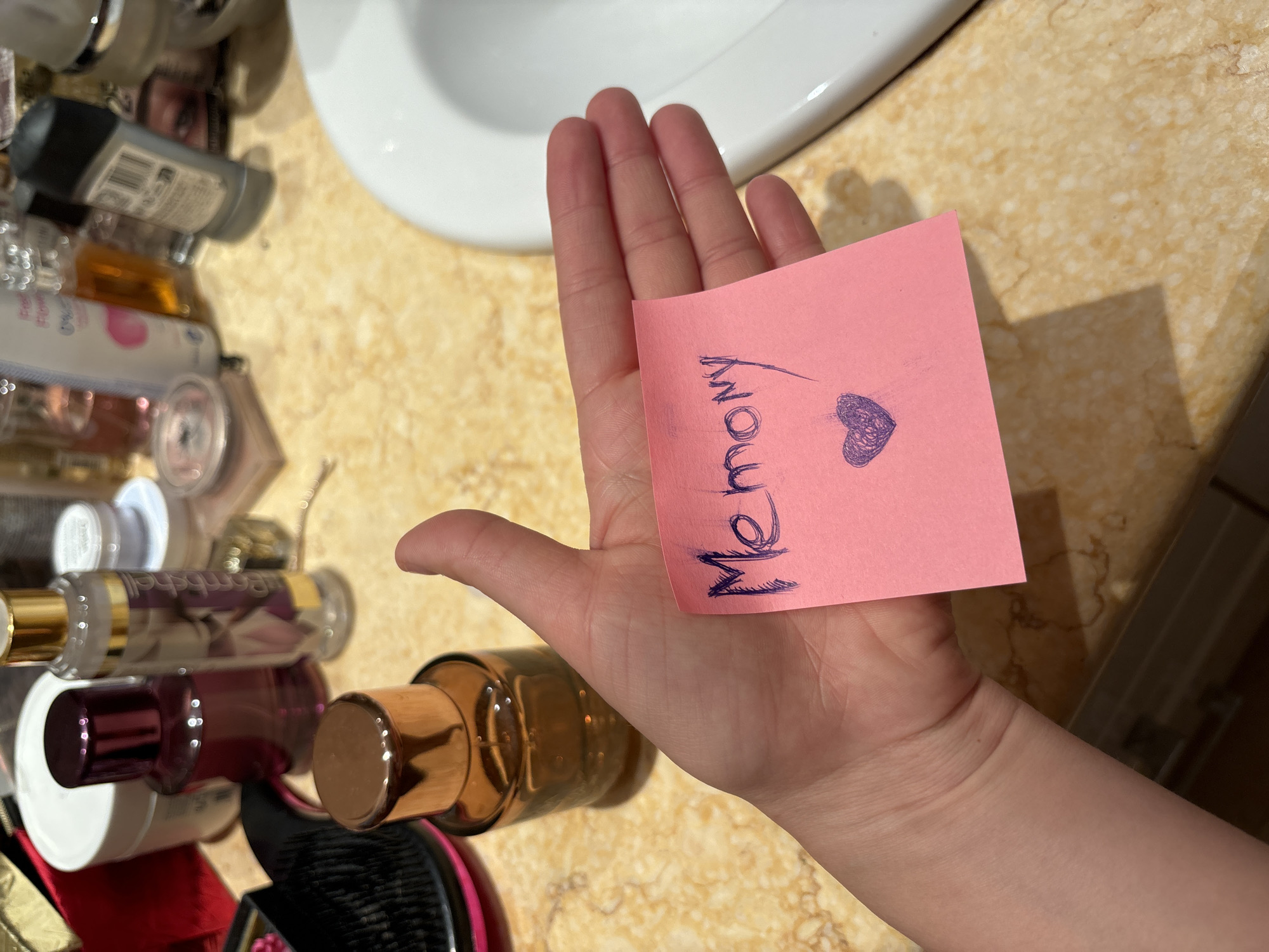An overhead, slightly angled photograph captures a human hand holding a pink rectangular sticky note with the palm facing upwards. The sticky note is positioned centrally in the frame, displaying the handwritten word "Memory" in dark blue or purple ink, followed by a shaded heart symbol in the same color directly beneath the text.
The background depicts a cluttered countertop, characteristic of a bathroom or vanity area. To the left of the hand, an amber-colored bottle with a metallic bronze/gold cap is partially visible in the lower-left foreground. Behind it, a dark red/burgundy cylindrical bottle and a clear bottle with a gold cap, bearing the vertical script label "Bombshell," are present. Further back on the countertop, a white cylindrical container with a lid, a smaller white jar, and a partially visible black hairbrush with pink accents are arranged.
To the right side of the image, a portion of a white ceramic sink basin is visible, along with a grey/black deodorant stick and other blurred bottles and personal care items. The countertop surface is a yellowish-brown material, featuring darker vein patterns consistent with marble or granite. The lighting indicates an indoor setting. The specific city of Budva, Montenegro, cannot be determined from the visual information presented in the image.